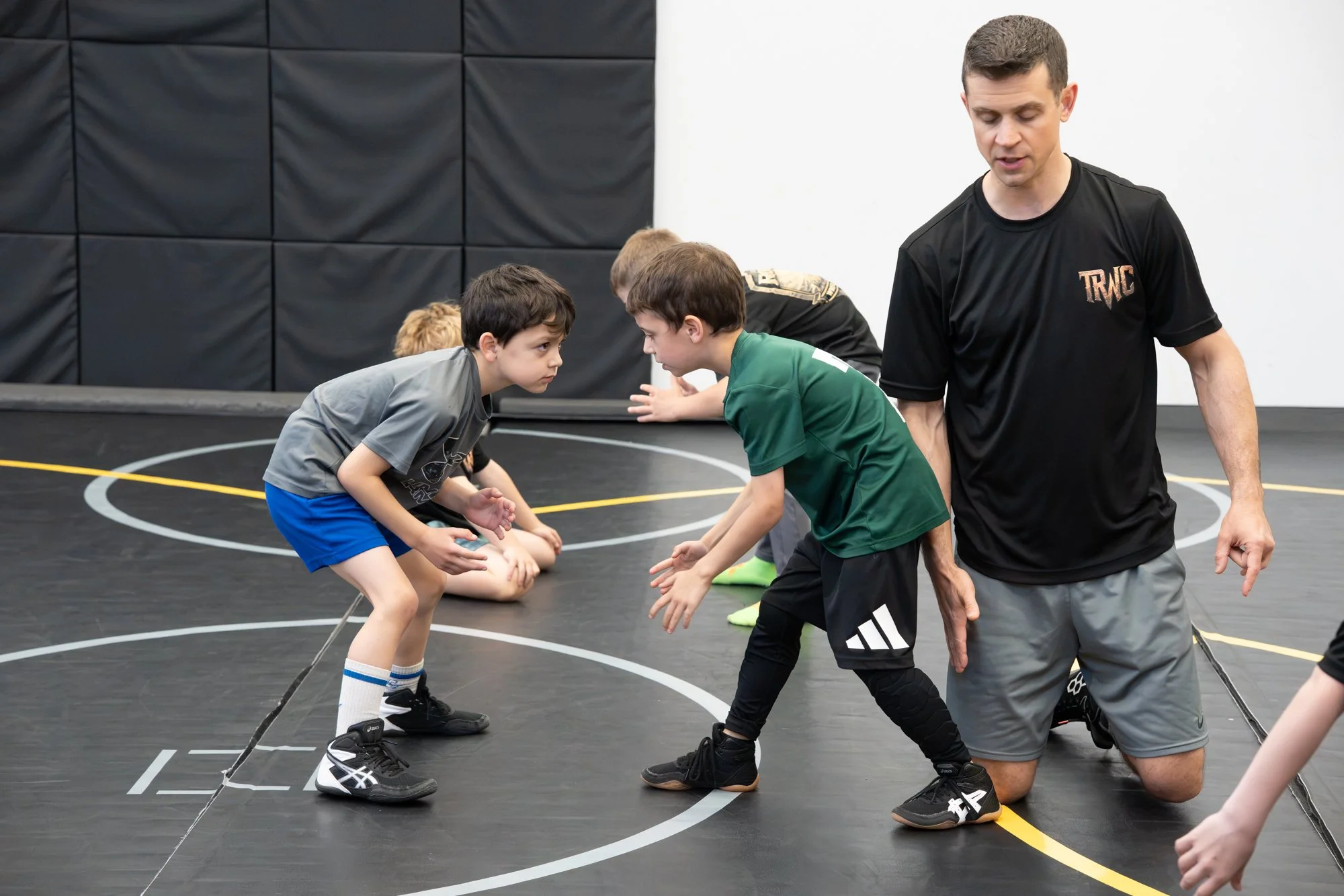 Two young boys facing each other on a wrestling mat, appearing to prepare for a match, with a coach guiding them