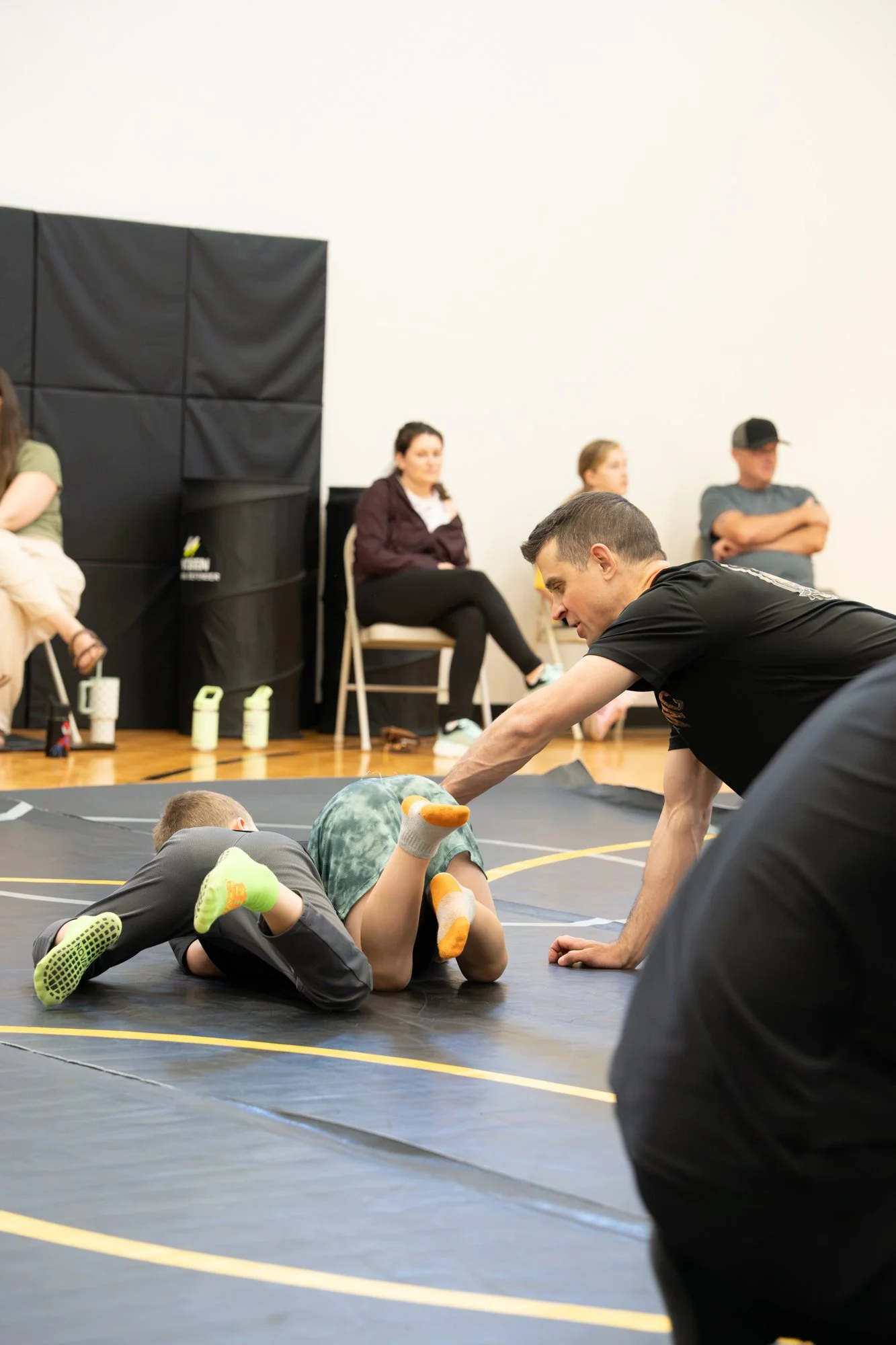 A wrestling practice or class with children practicing on a mat with instructors assisting. Children are dressed in game costumes, and some are in a wrestling stance while others are lying face down. In the background, four people are sitting on chai