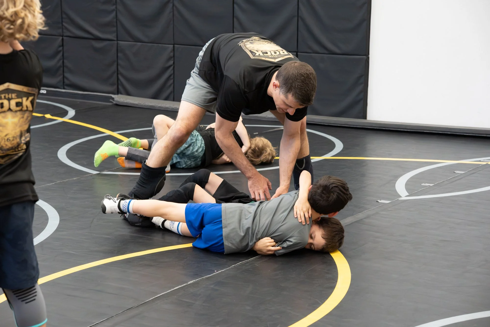 A wrestling coach helps a group of young wrestlers practice on a black wrestling mat in a gym, with padded walls in the background.