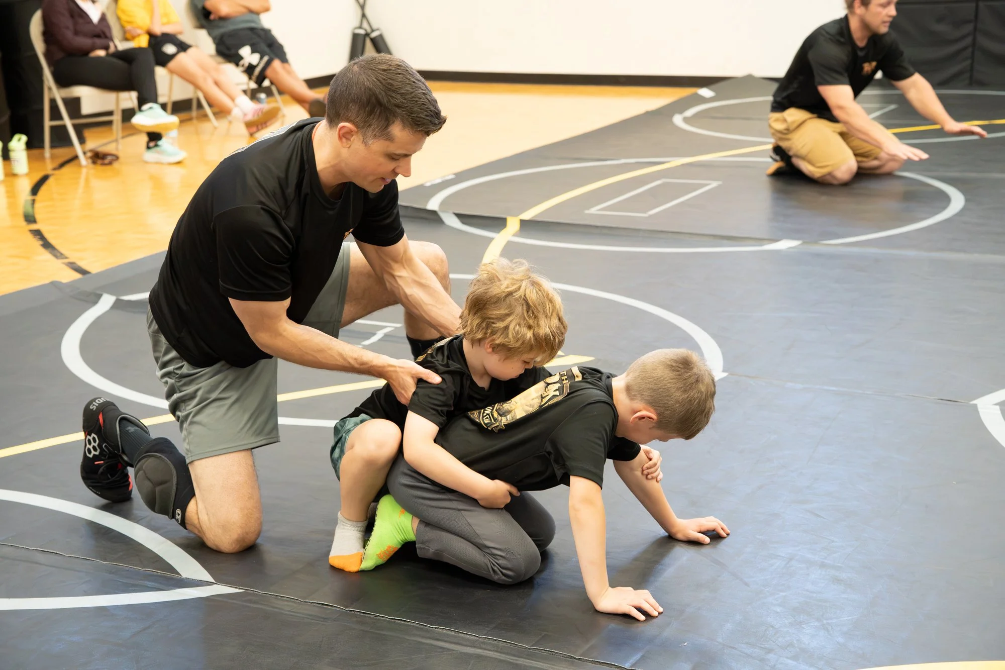 A wrestling coach helping young wrestlers with a warm-up or stretching exercises on a black wrestling mat in a gymnasium. Four kids are lying on their stomachs, while they do push-ups or stretching, with the coach assisting. Three people are sitting 