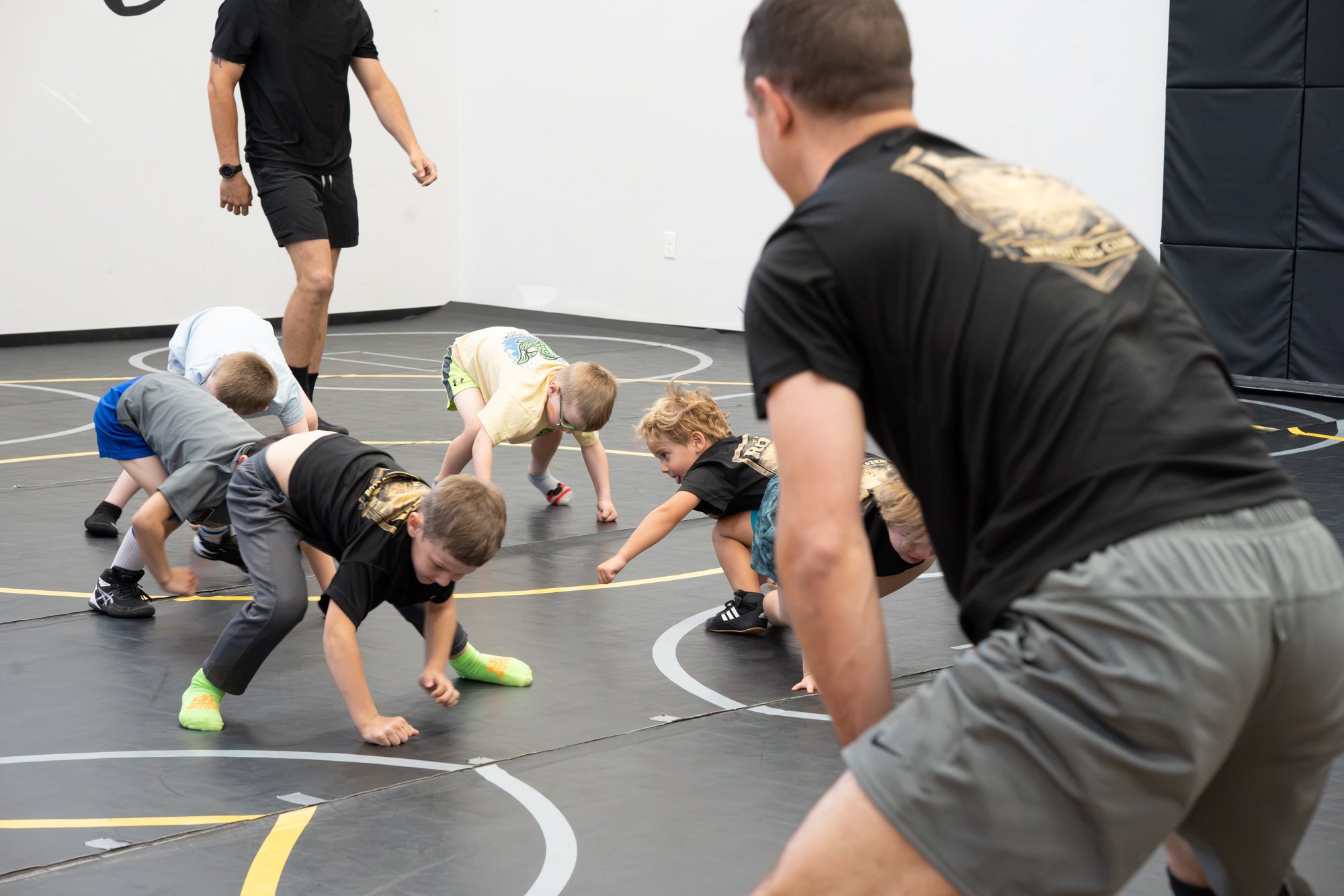 Children participating in a wrestling drill on a black indoor mat, with two adult supervisors overseeing the activity.