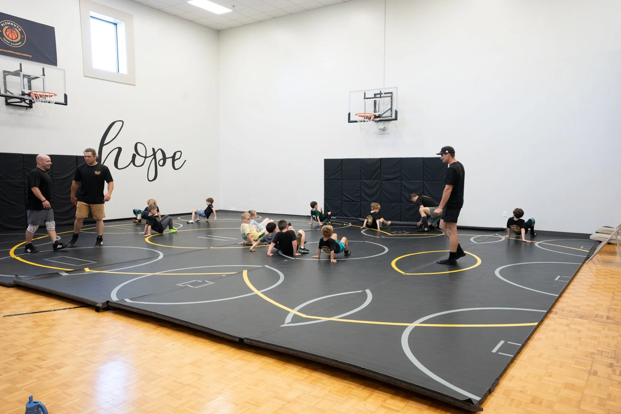 Children and coaches in a gymnasium with a wrestling mat during a wrestling class or practice session.