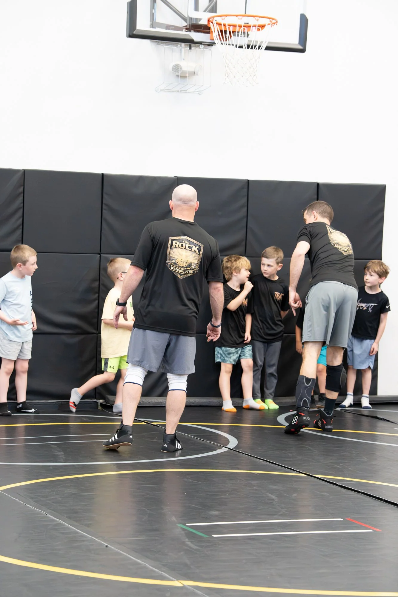 Boys in a gymnasium participating in a wrestling or sports practice, with two coaches or trainers instructing them.