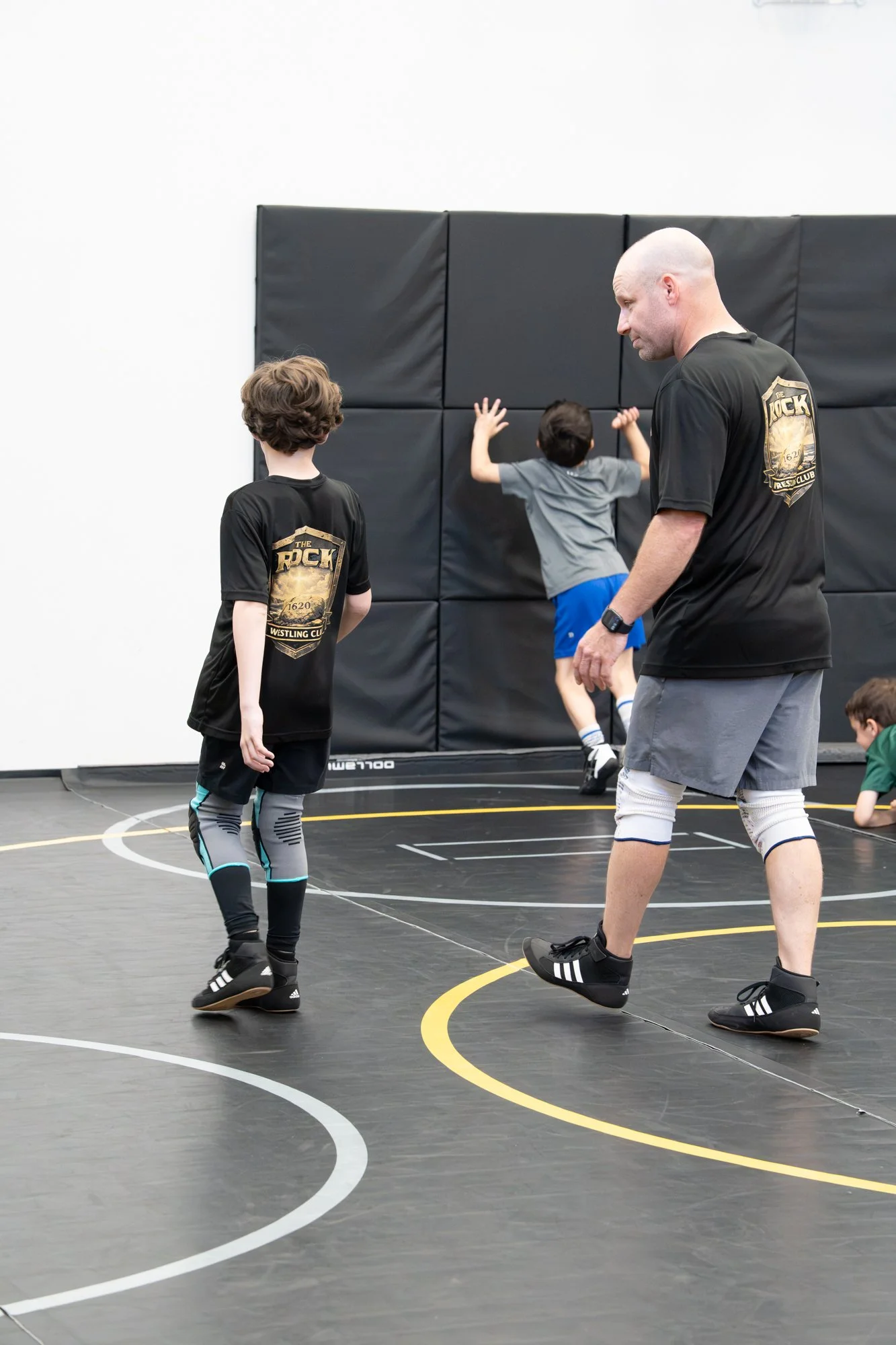 Indoor wrestling practice with children and coach. Children are warming up or stretching on wrestling mat, coach observing. Black padded wall in background.