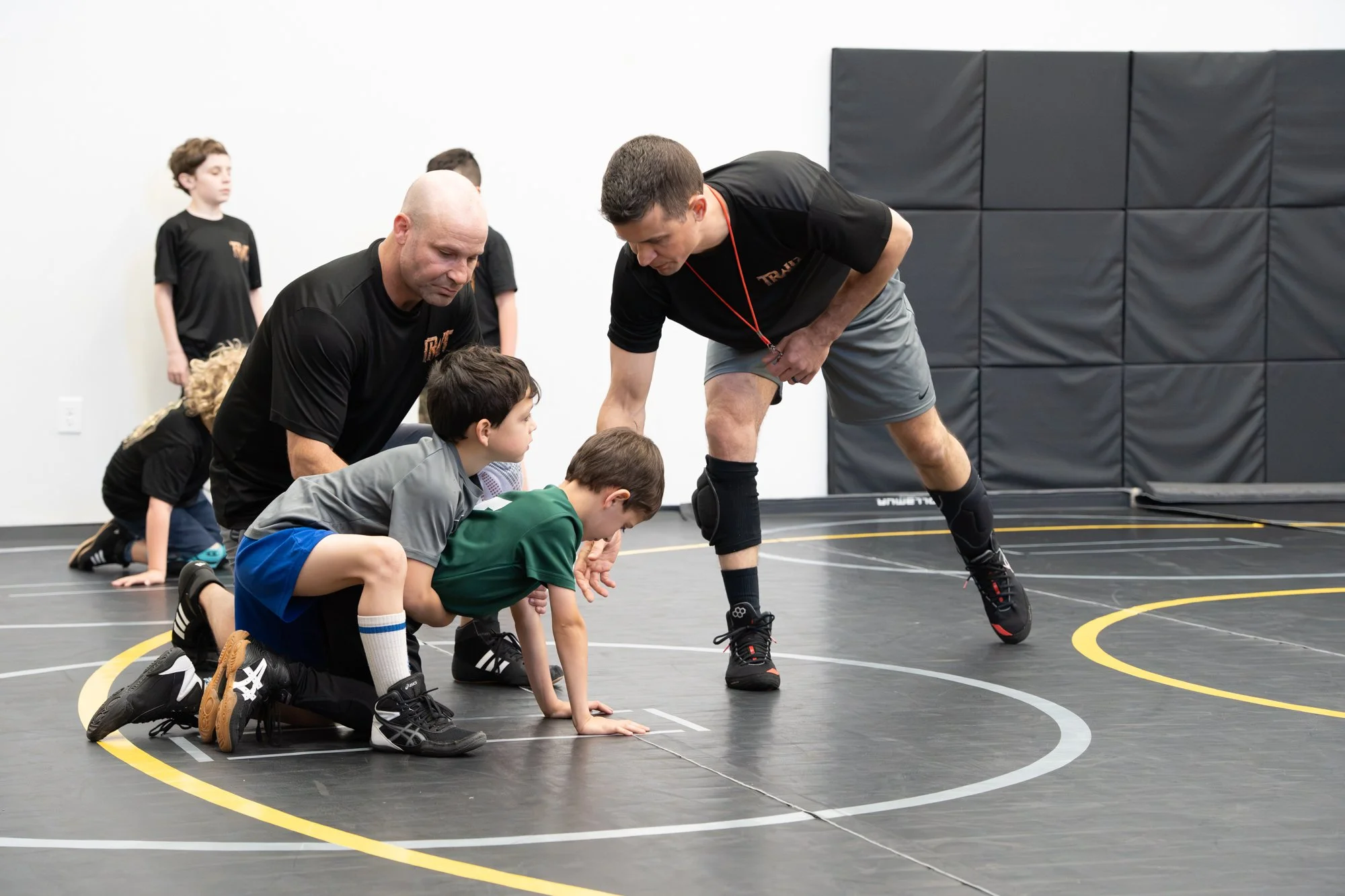 Children and coaches in a wrestling gym practicing a drill on a black wrestling mat with yellow and white lines, black padded walls, and a plain white wall in the background.