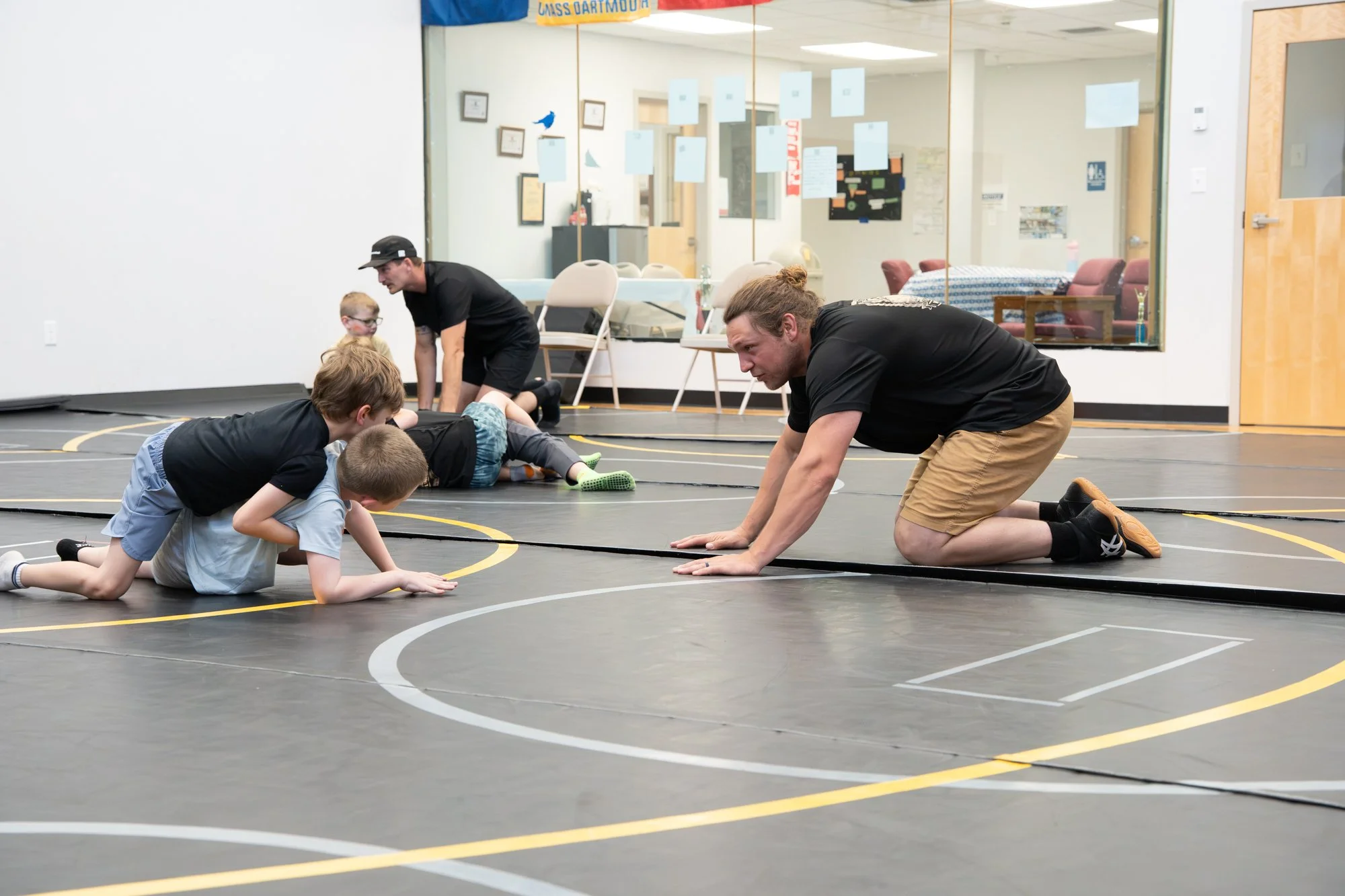 Three children and two adults practicing wrestling or a similar activity on a gymnasium floor with wrestling mats. The adults are helping the children, who are on their hands and knees, focused on their activity, in a room with chairs, framed picture