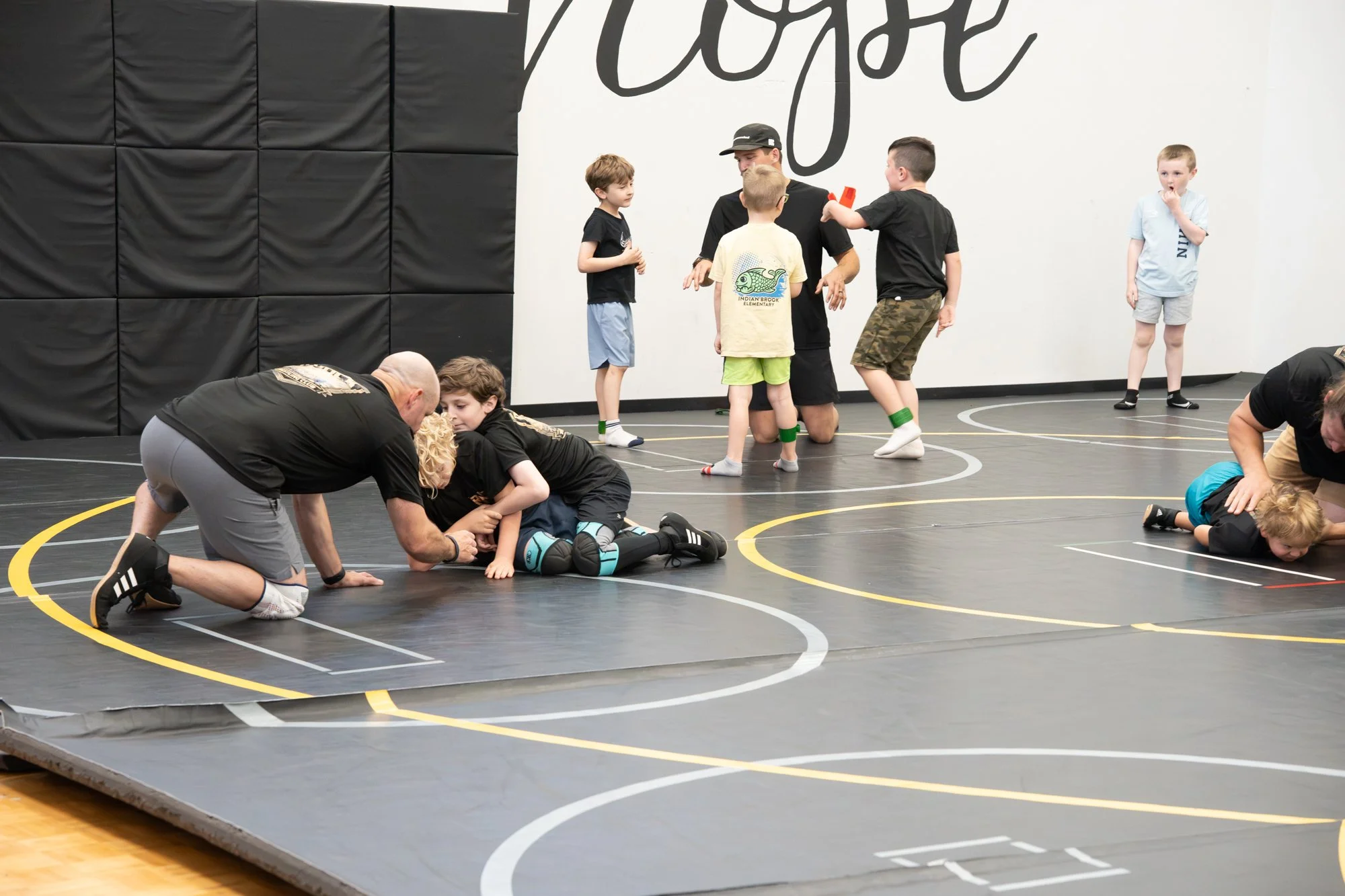 Children and adults practicing wrestling or grappling on a black wrestling mat inside a gymnasium, with some children standing and others lying or being assisted on the mat.