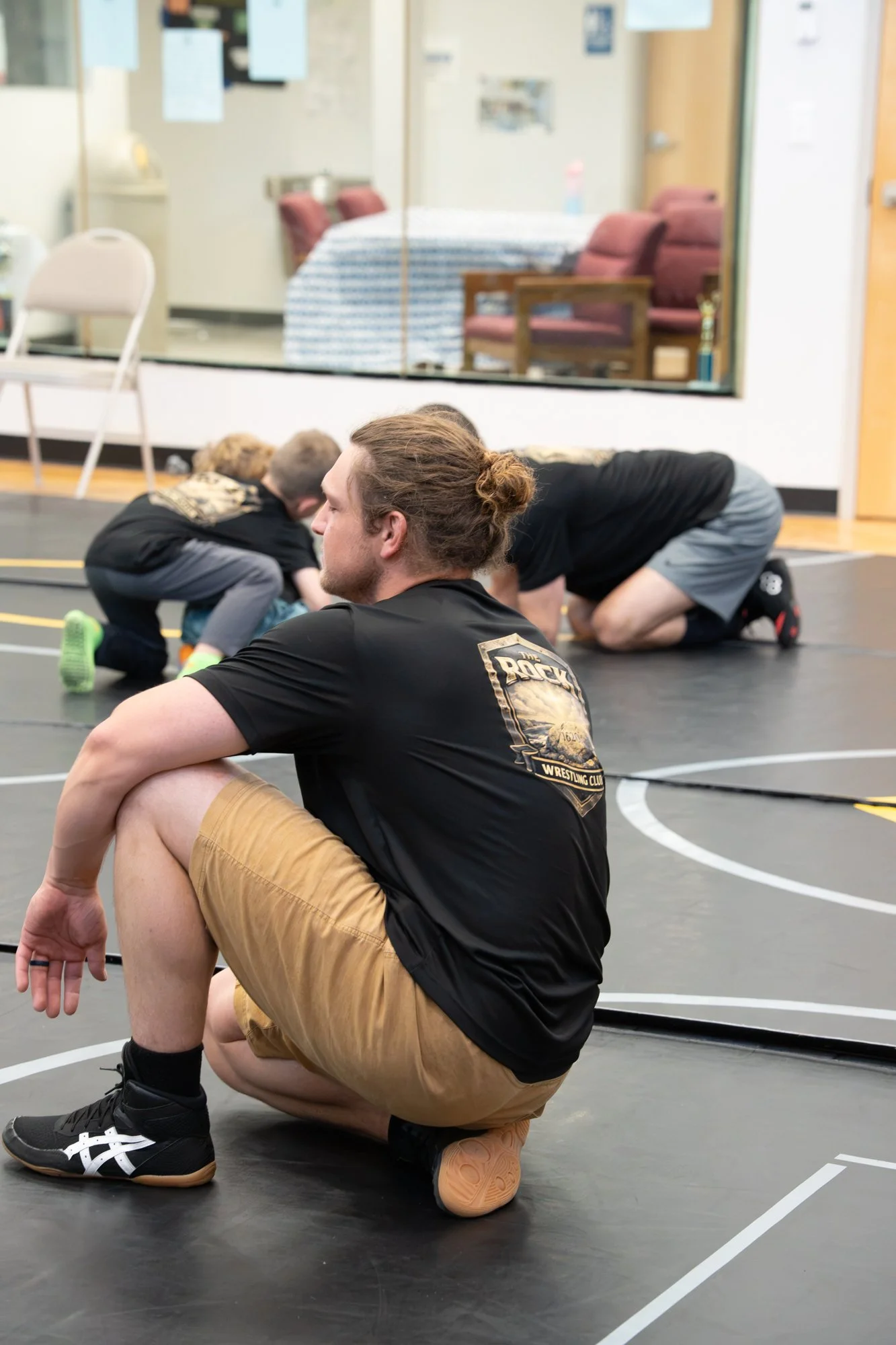A man and a child practicing wrestling on a gym mat, with a glass partition showing a room with furniture in the background.