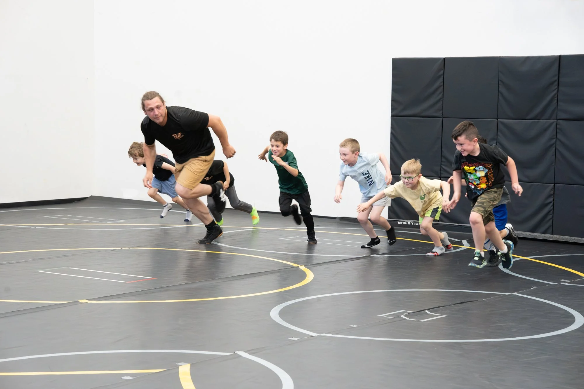 A group of children and an adult participating in a running activity in an indoor gym. The kids are wearing casual athletic clothes and running on a marked sports court with black wall padding in the background.