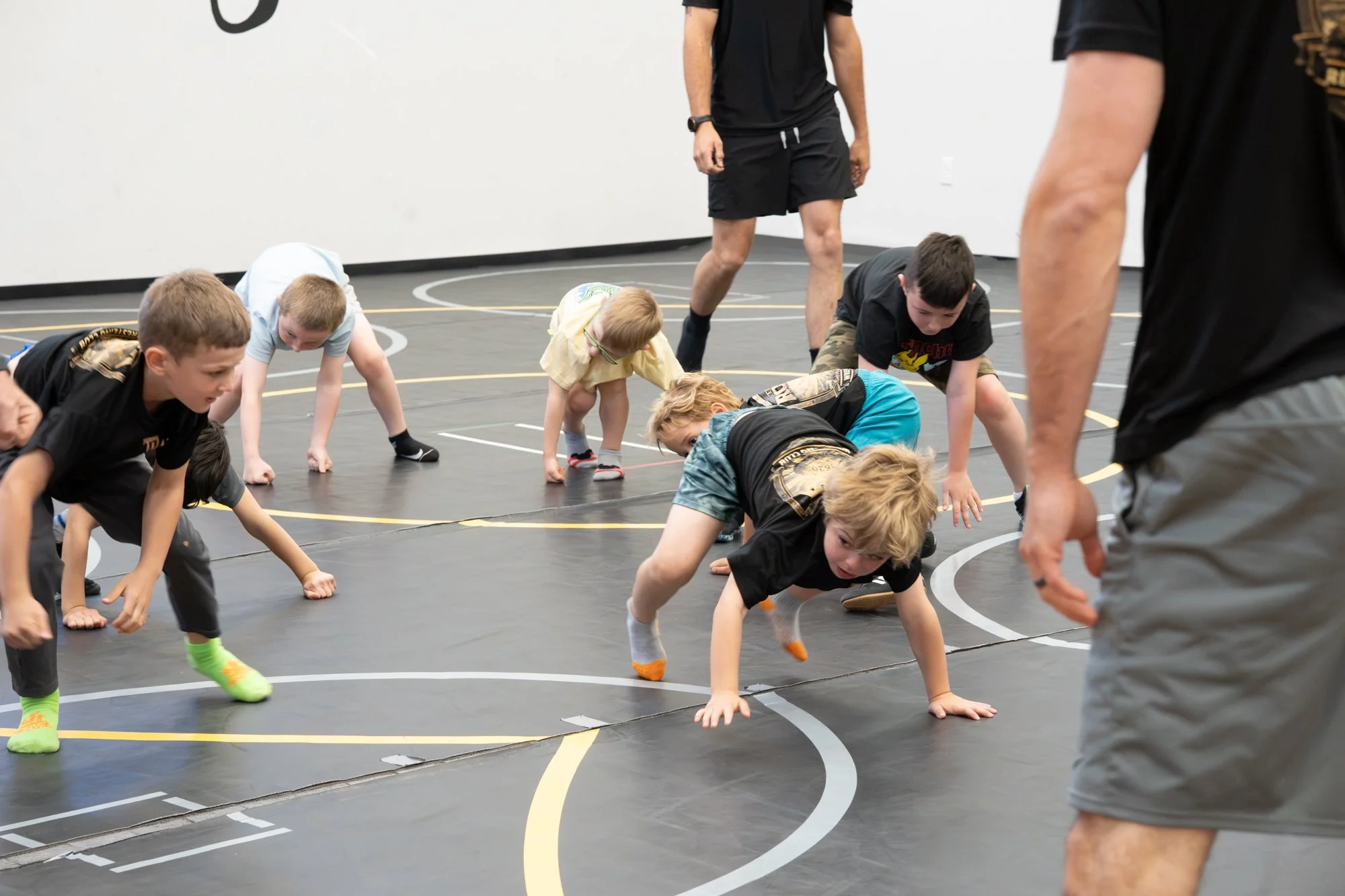 Children participating in a wrestling drill on a gymnasium floor, with coaches guiding them.