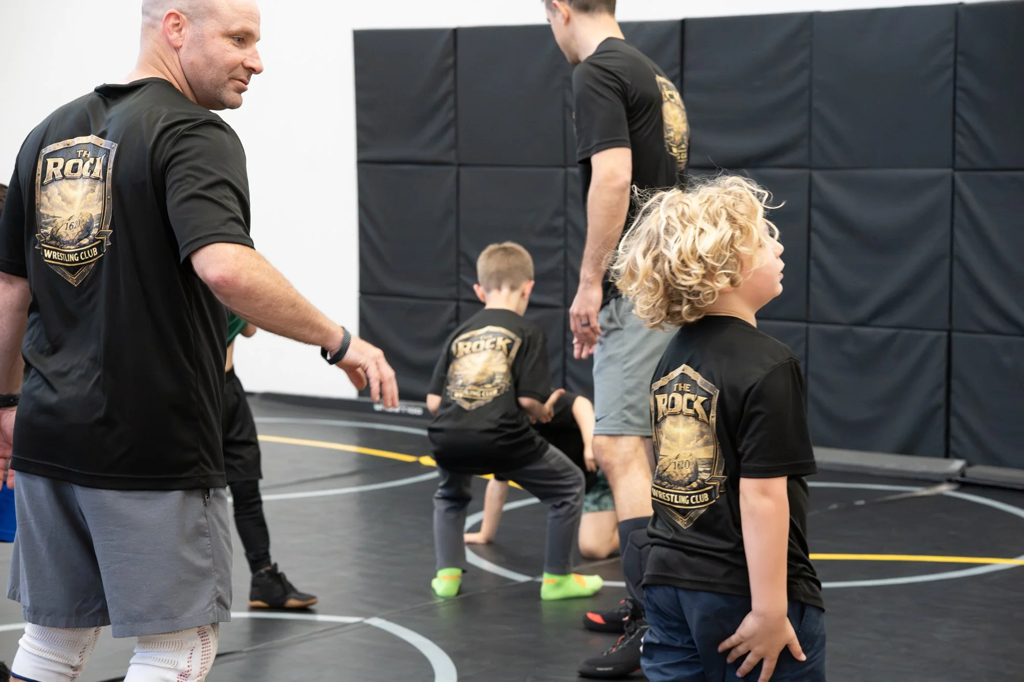 Wrestlers in black shirts with 'The Rock Wrestling Club' logo participating in a wrestling practice, with one adult male instructor and several young children on a black wrestling mat with black padded walls.