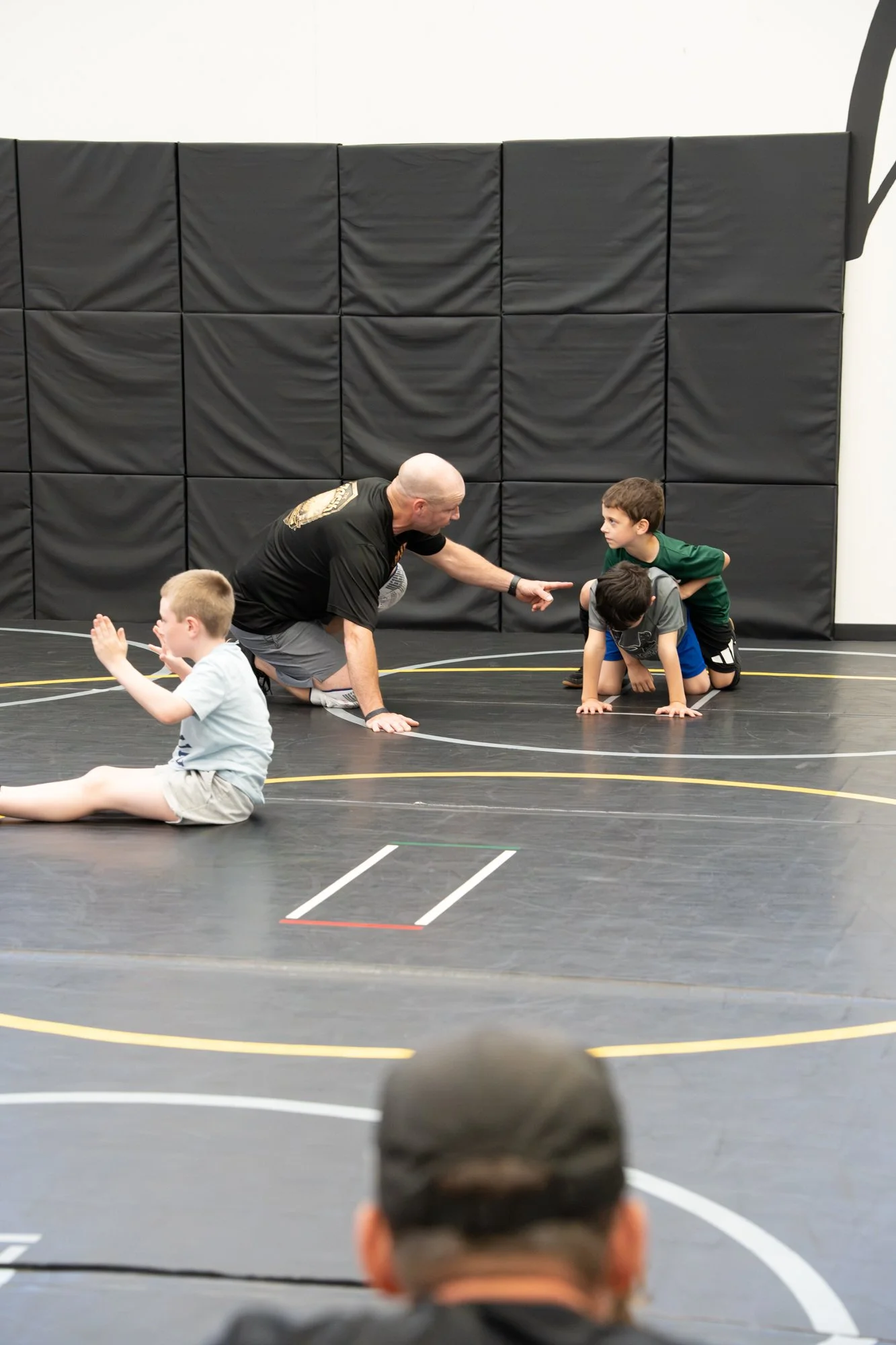 A wrestling coach instructs a young boy in a wrestling gym, with other children, on a wrestling mat with black padding and wall padding in the background.