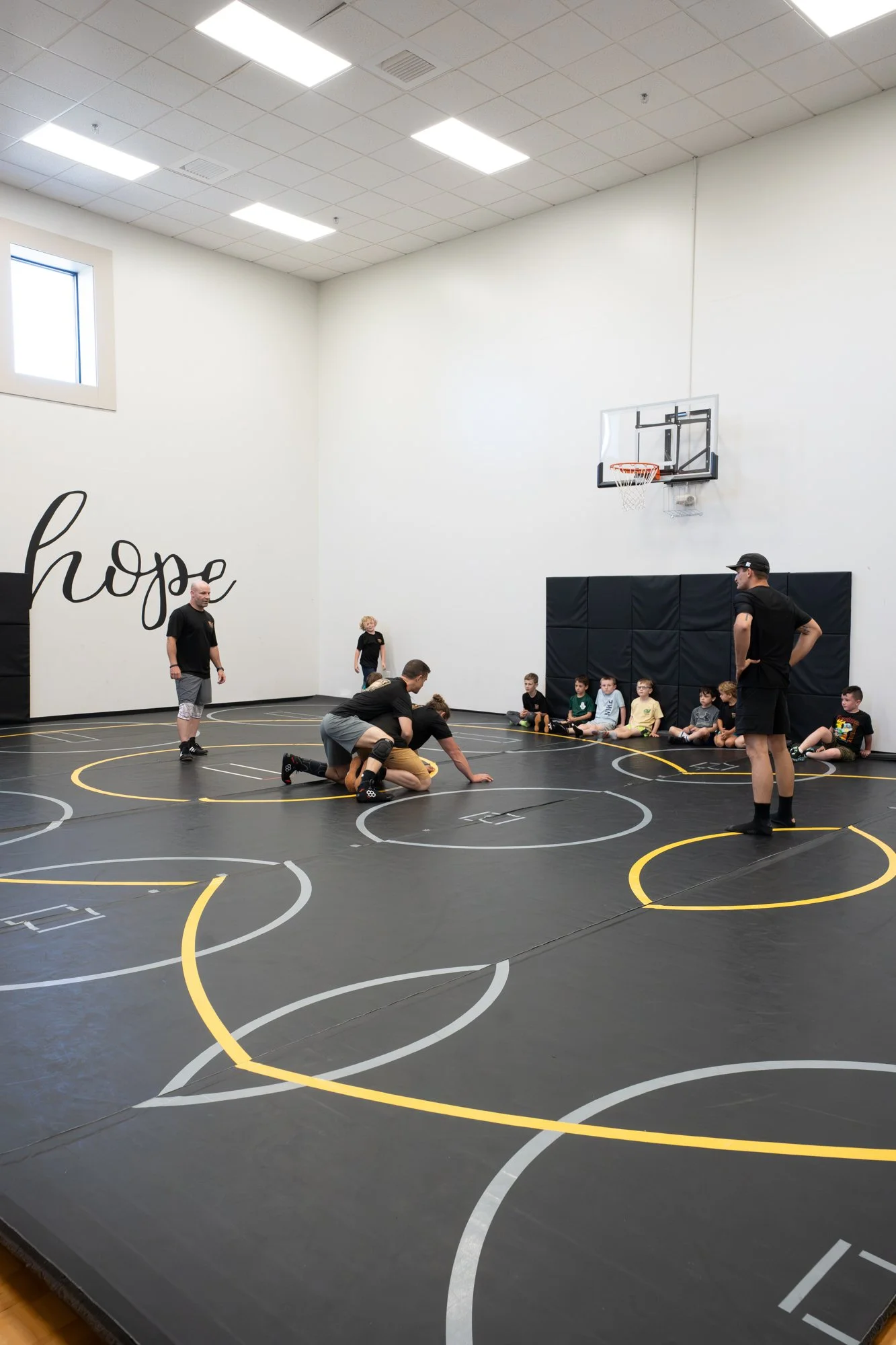 A gymnasium with a wrestling or martial arts training mat, where a group of children and instructors are practicing or participating in a class. The mat has circular markings and a wall decorated with the word 'hope' in large cursive letters. There i