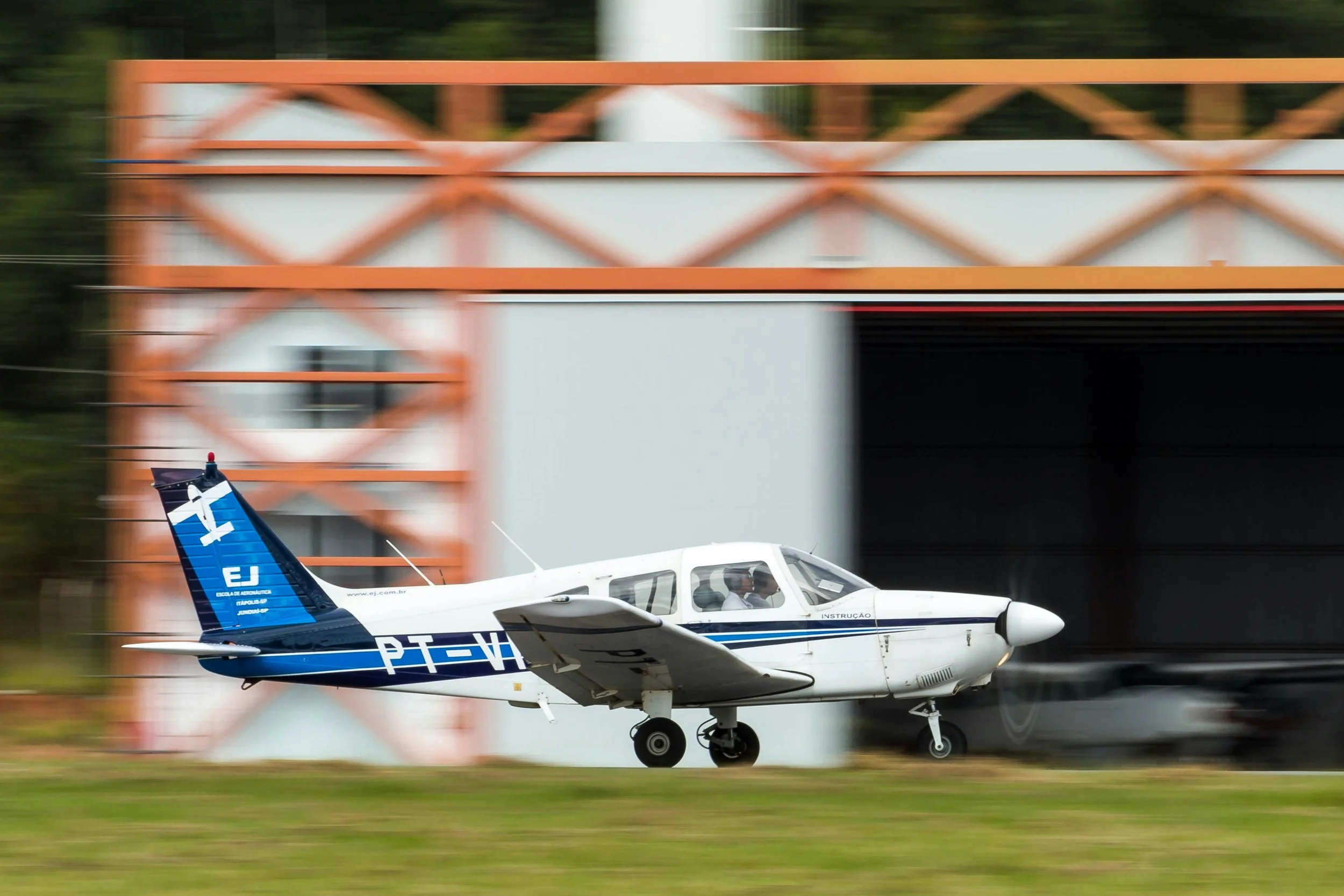 A small white and blue aircraft with the registration PT-HJH and the word 'Instrucao' on the side, taking off from an airfield with blurring of the background indicating motion.