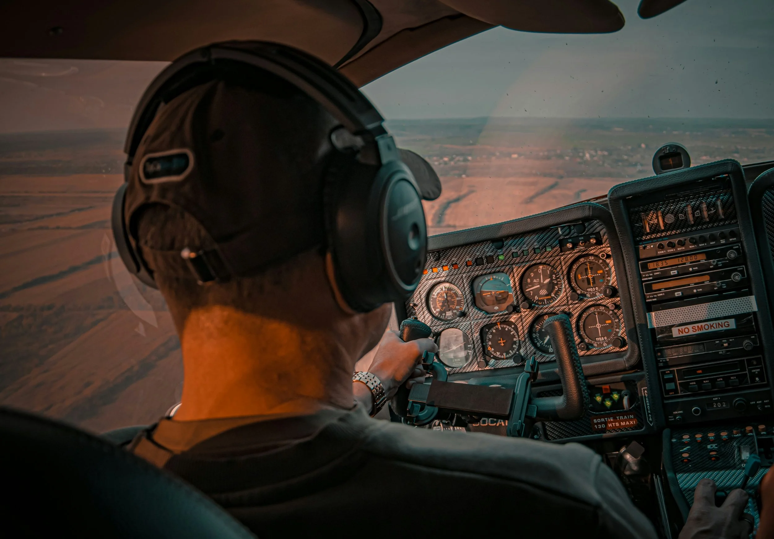 Pilot in the cockpit of an aircraft, flying over a landscape during daylight, wearing a headset.