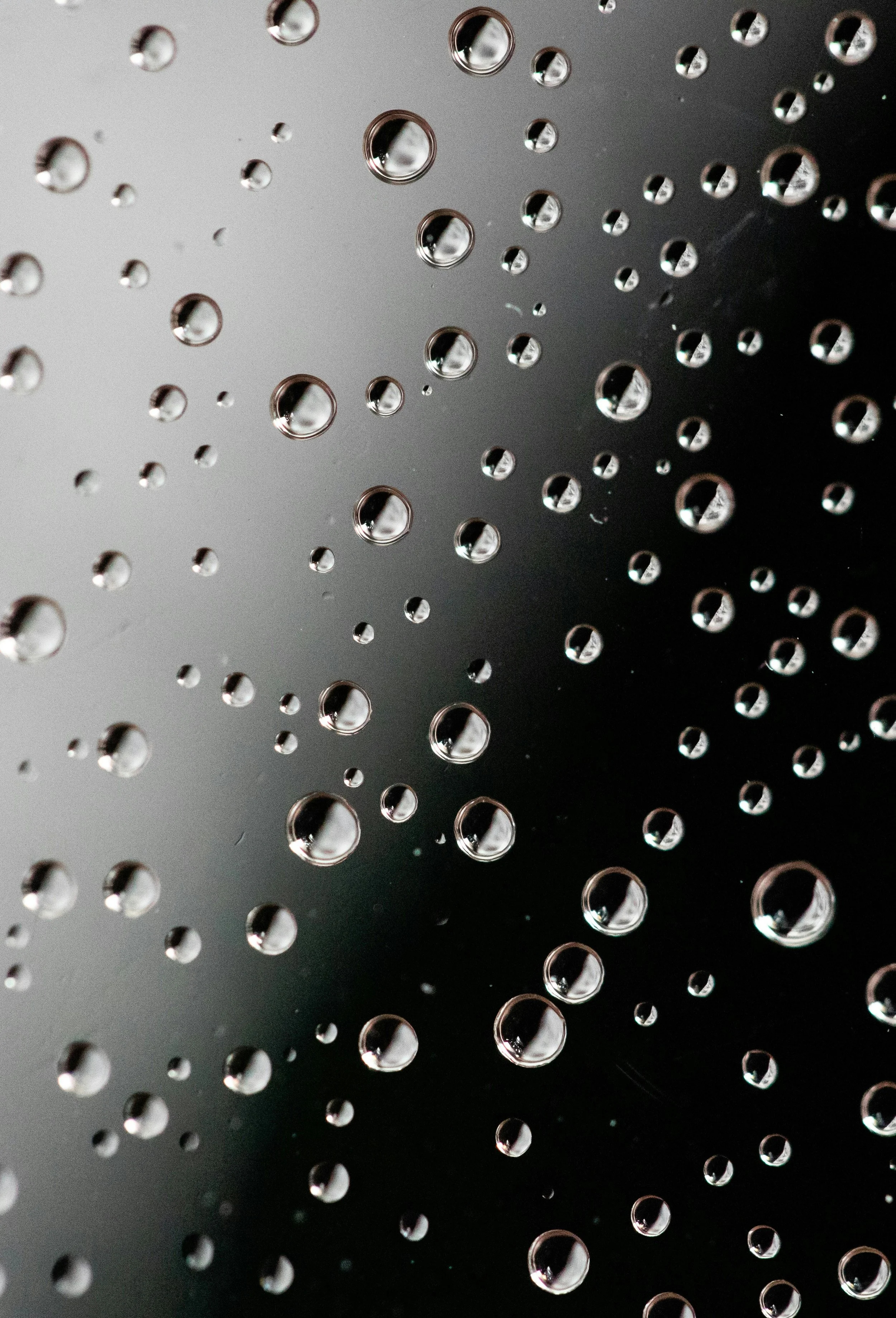 Close-up of water droplets on a smooth, shiny surface, possibly glass or metal, with reflections and varying sizes, against a dark background.