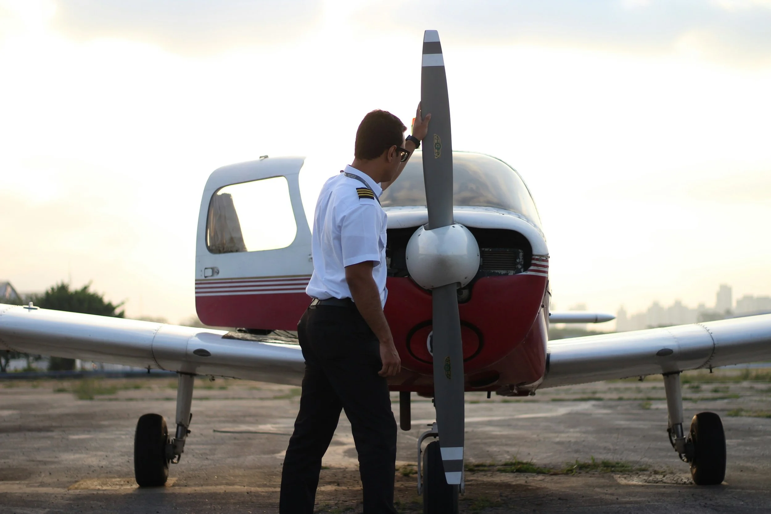 A pilot in uniform standing next to a small white and red airplane, touching a vertical stabilizer on the aircraft's tail, with a city skyline in the distance during sunset.