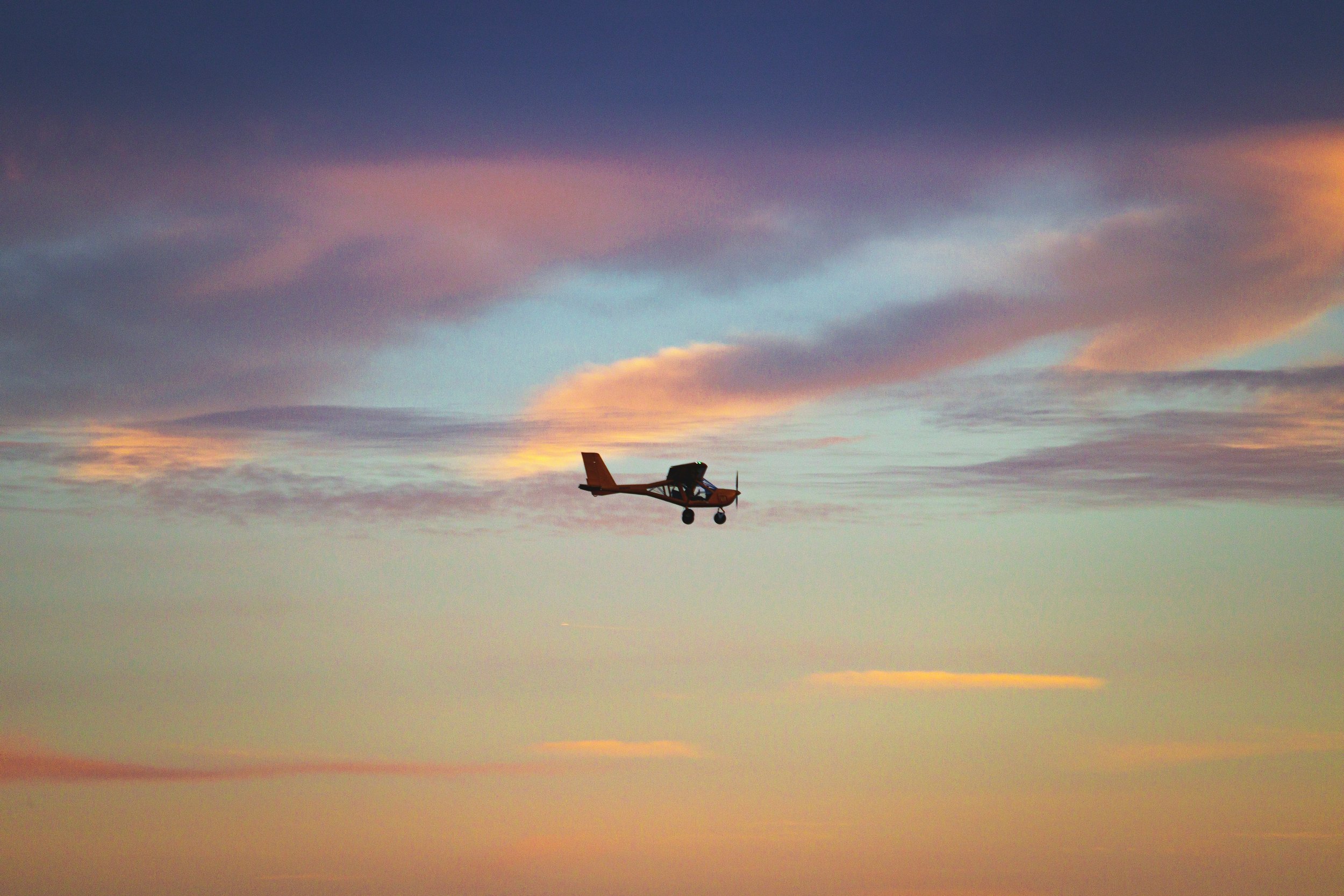 Small orange airplane flying in a colorful sky with pink, purple, and blue clouds at sunset or sunrise.