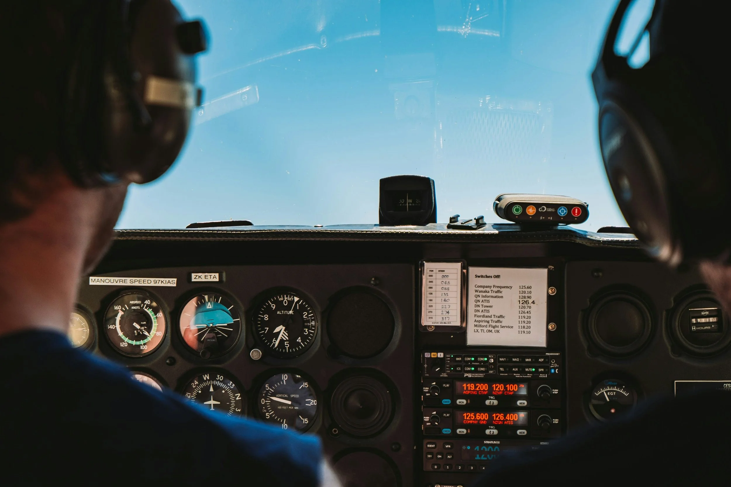 View from inside a cockpit airplane showing navigation instruments, radios, and a digital display with frequency information, with a clear blue sky through the windshield and two pilots visible on either side.