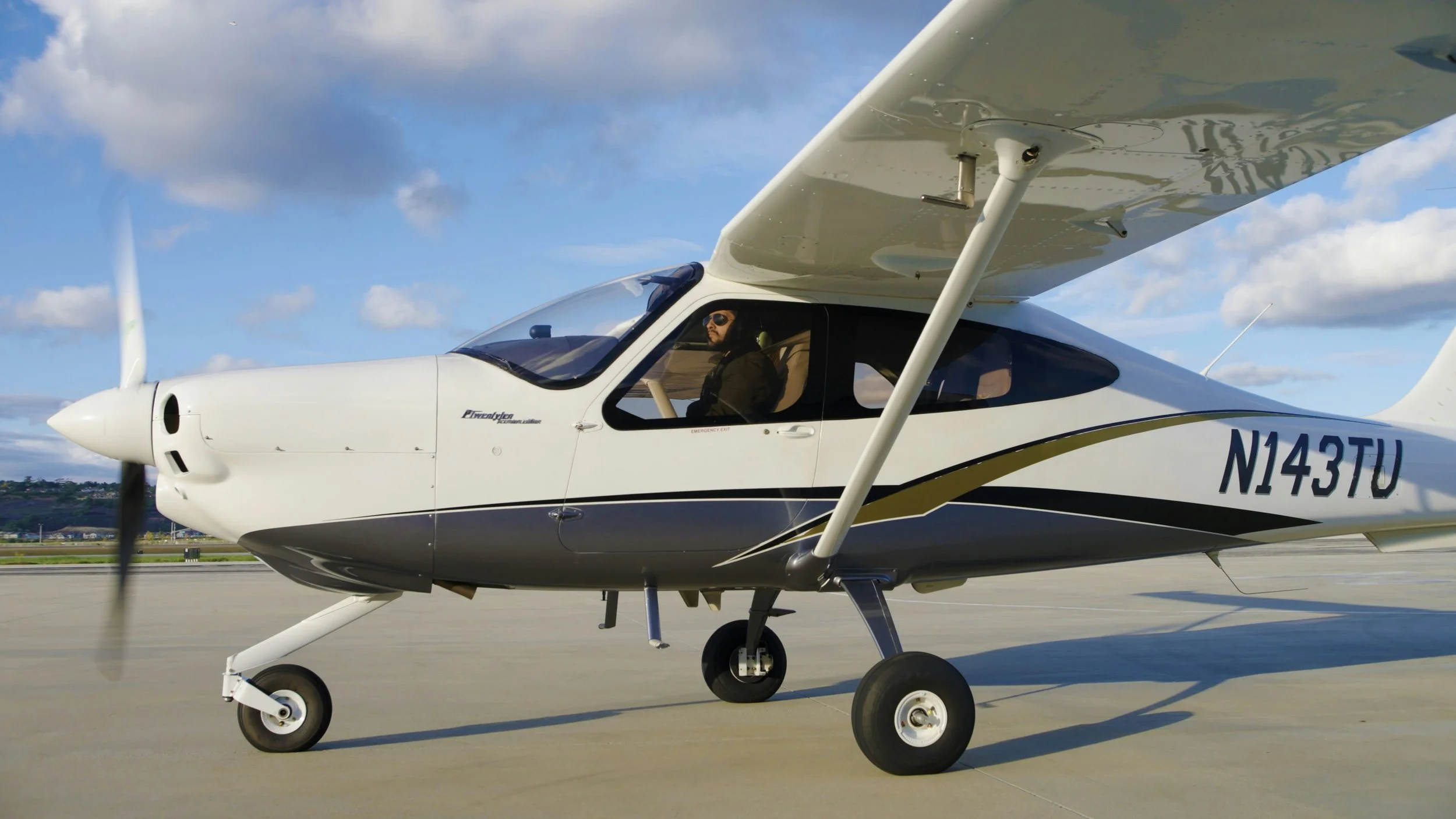 A small white and black propeller airplane with a pilot sitting inside, parked on an airport runway under a partly cloudy sky.