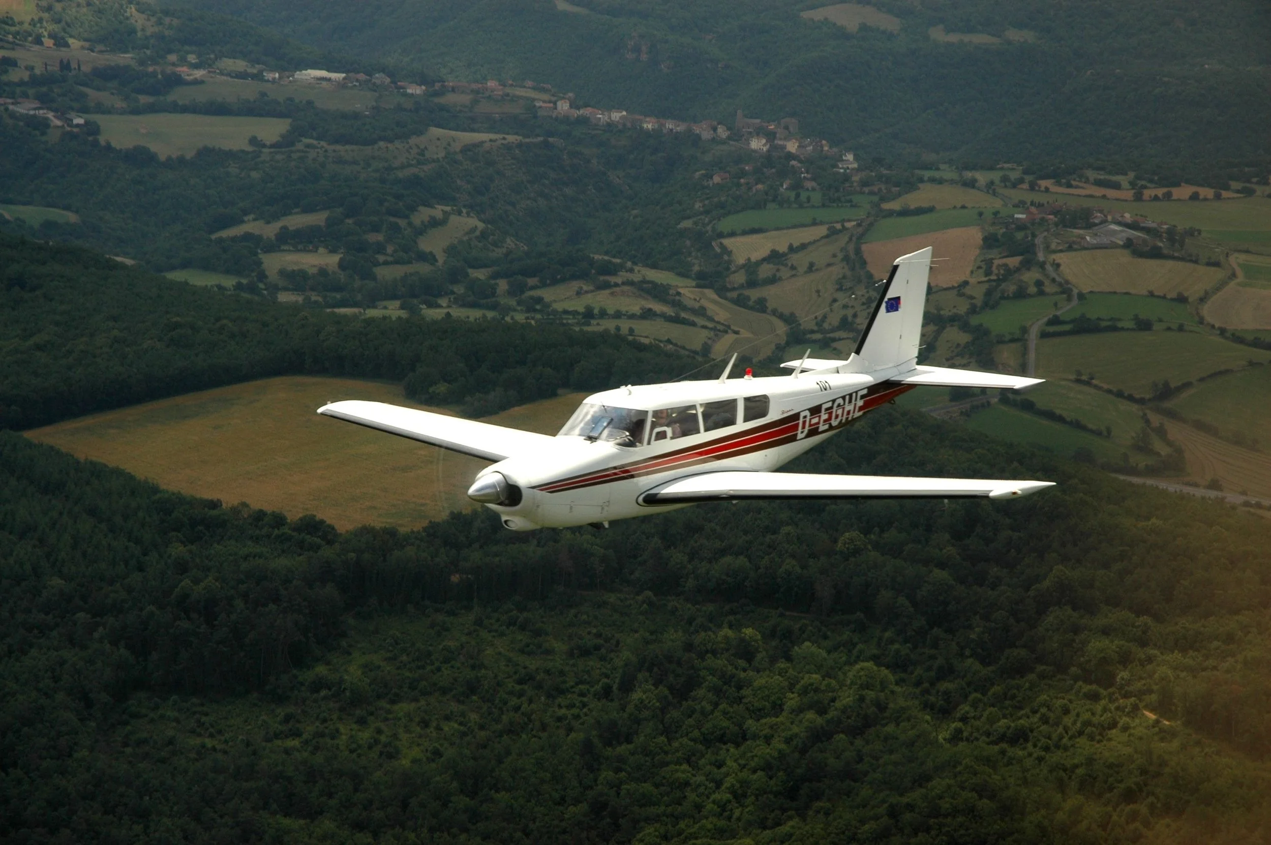 Small white airplane flying over green countryside hills and fields.