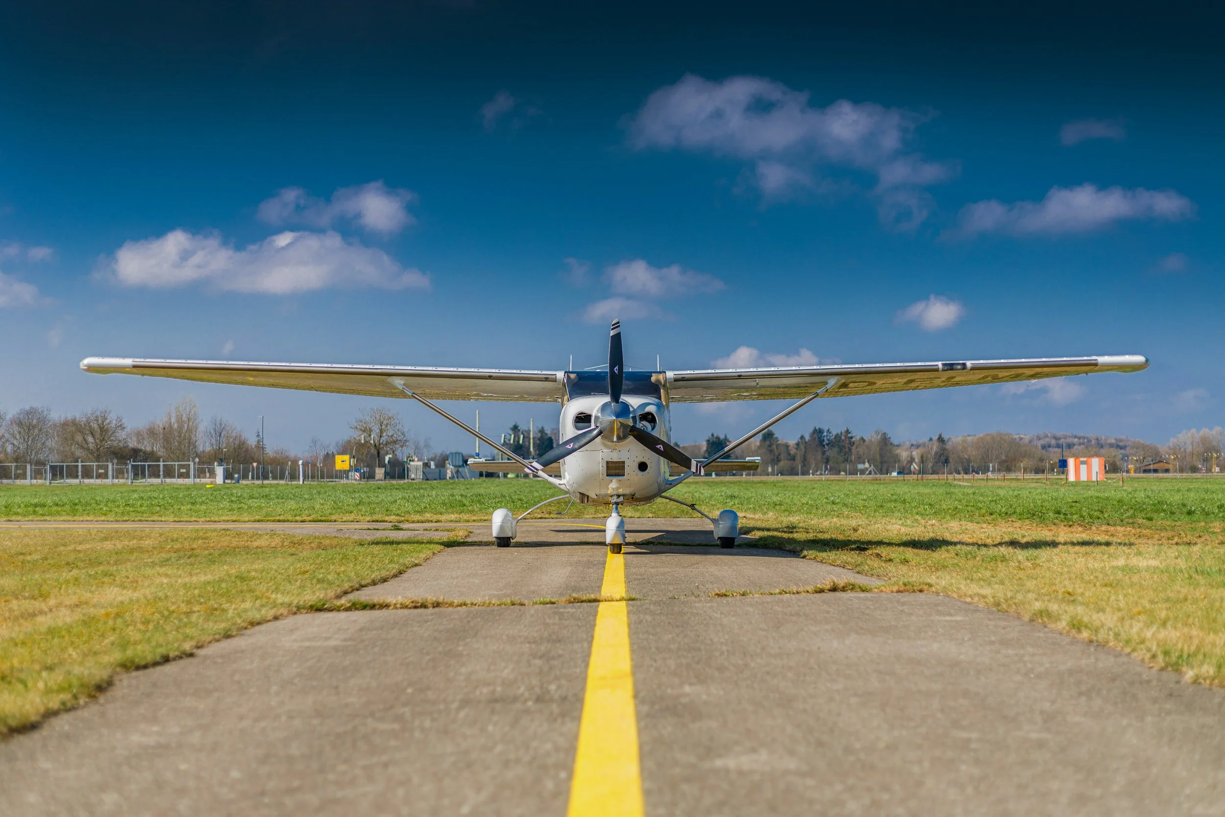 Front view of a small white airplane with blue accents on a tarmac, aligned on a yellow centerline, with a background of a grassy field and blue sky with clouds.
