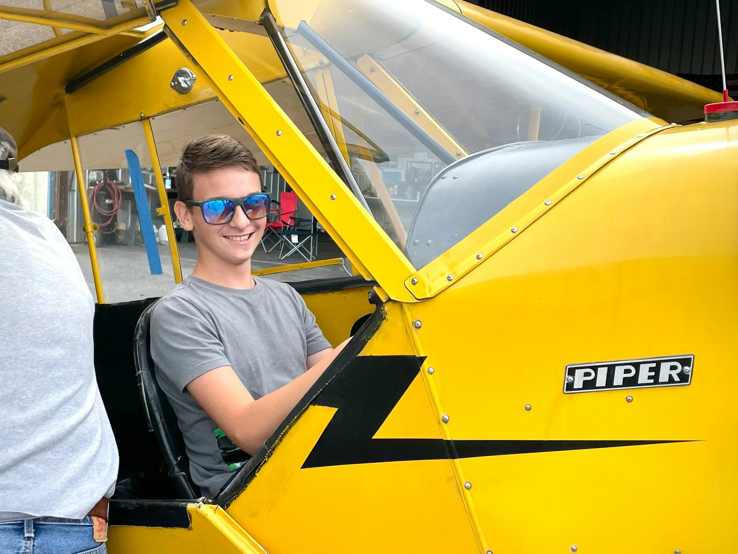 A young man wearing glasses and a gray T-shirt sitting in a yellow and black ultralight aircraft. The aircraft has a clear canopy and a sign that reads 'PIPER' on the side.