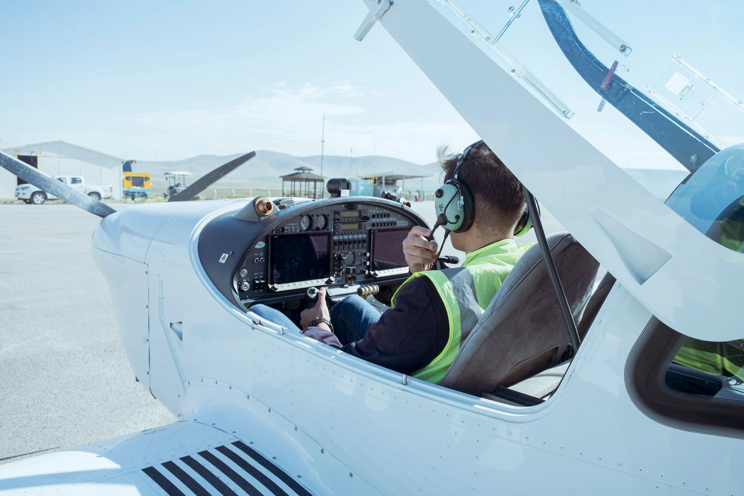 A person sitting inside the cockpit of a small white airplane at an airport, wearing headphones and a neon green safety vest, with open aircraft doors and airport structures in the background.
