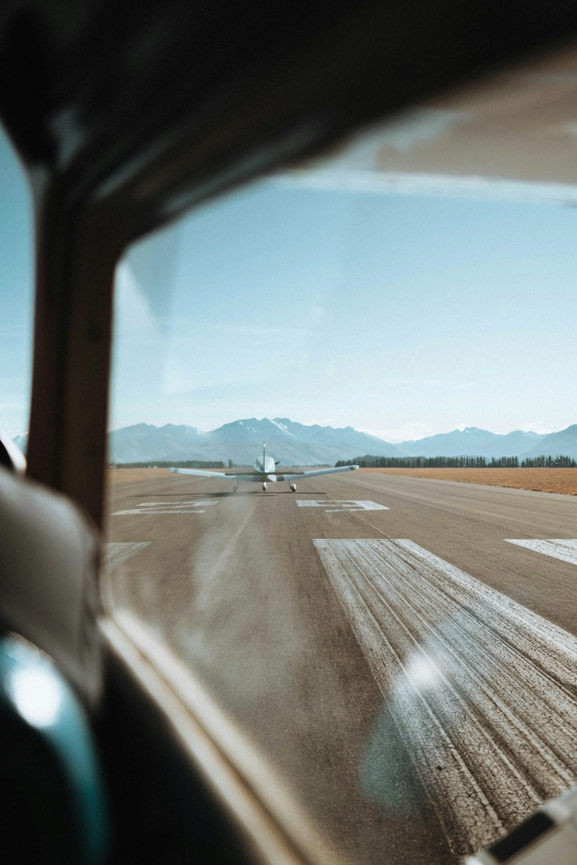Inside an aircraft cockpit with a view of the runway and a small airplane ahead, mountains in the background, and blue sky