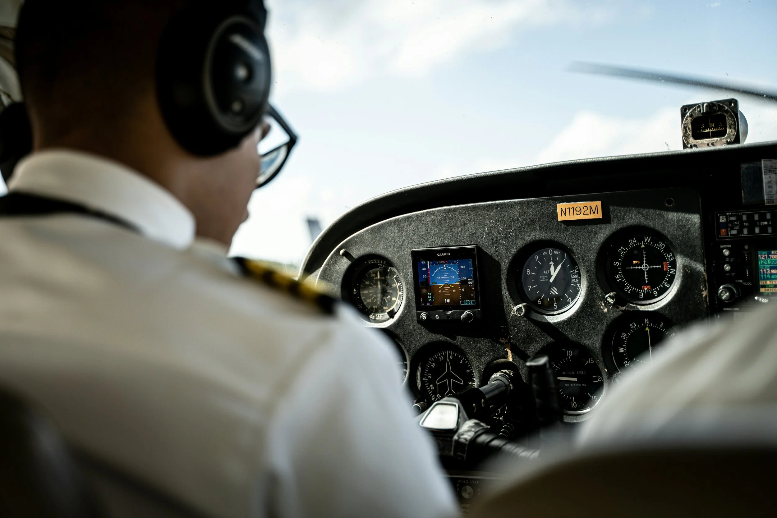 Close-up view of a pilot in cockpit wearing glasses and headphones, with various aircraft instruments and navigation equipment visible on the dashboard.
