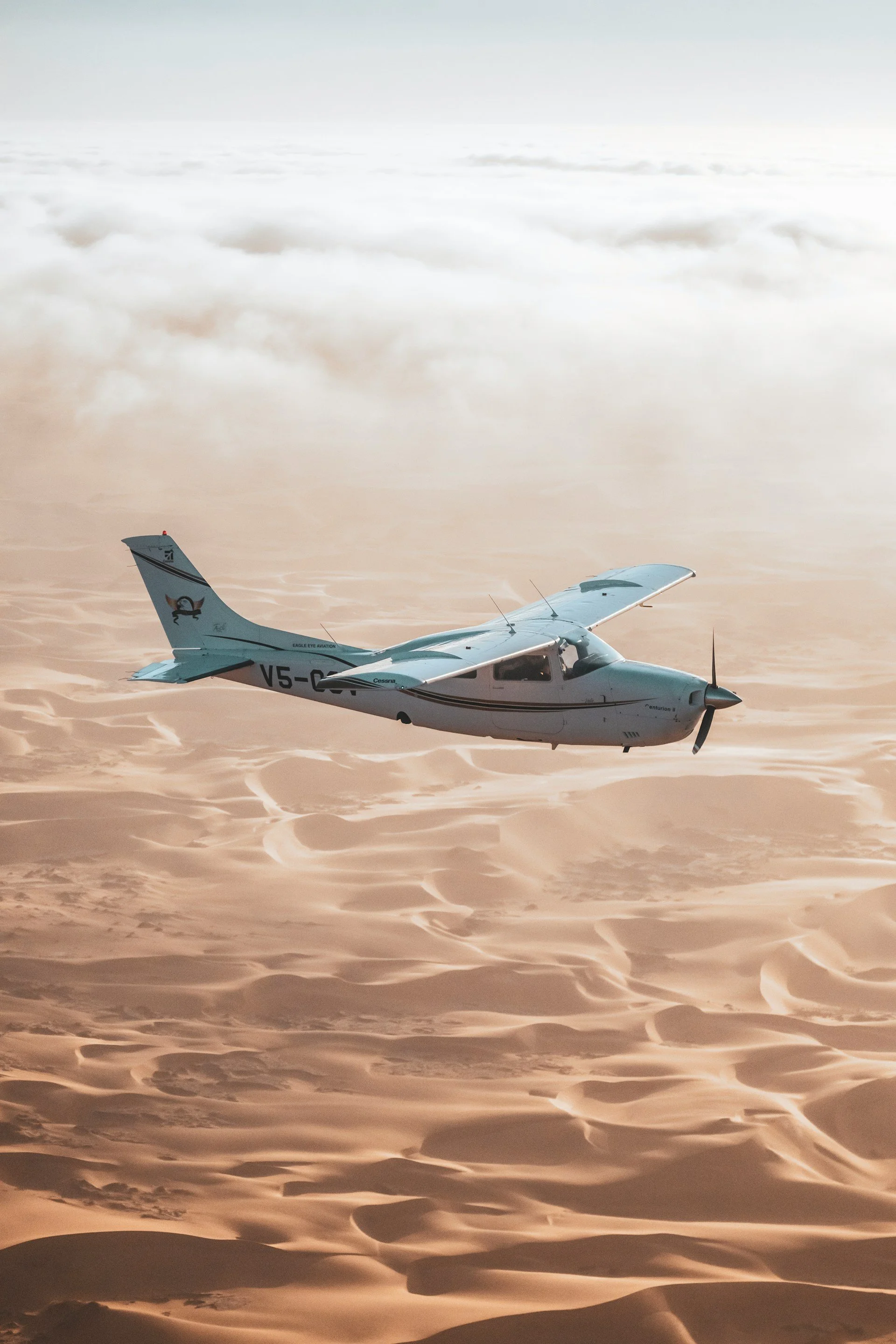 Small white airplane flying above desert sand dunes at sunset or sunrise.