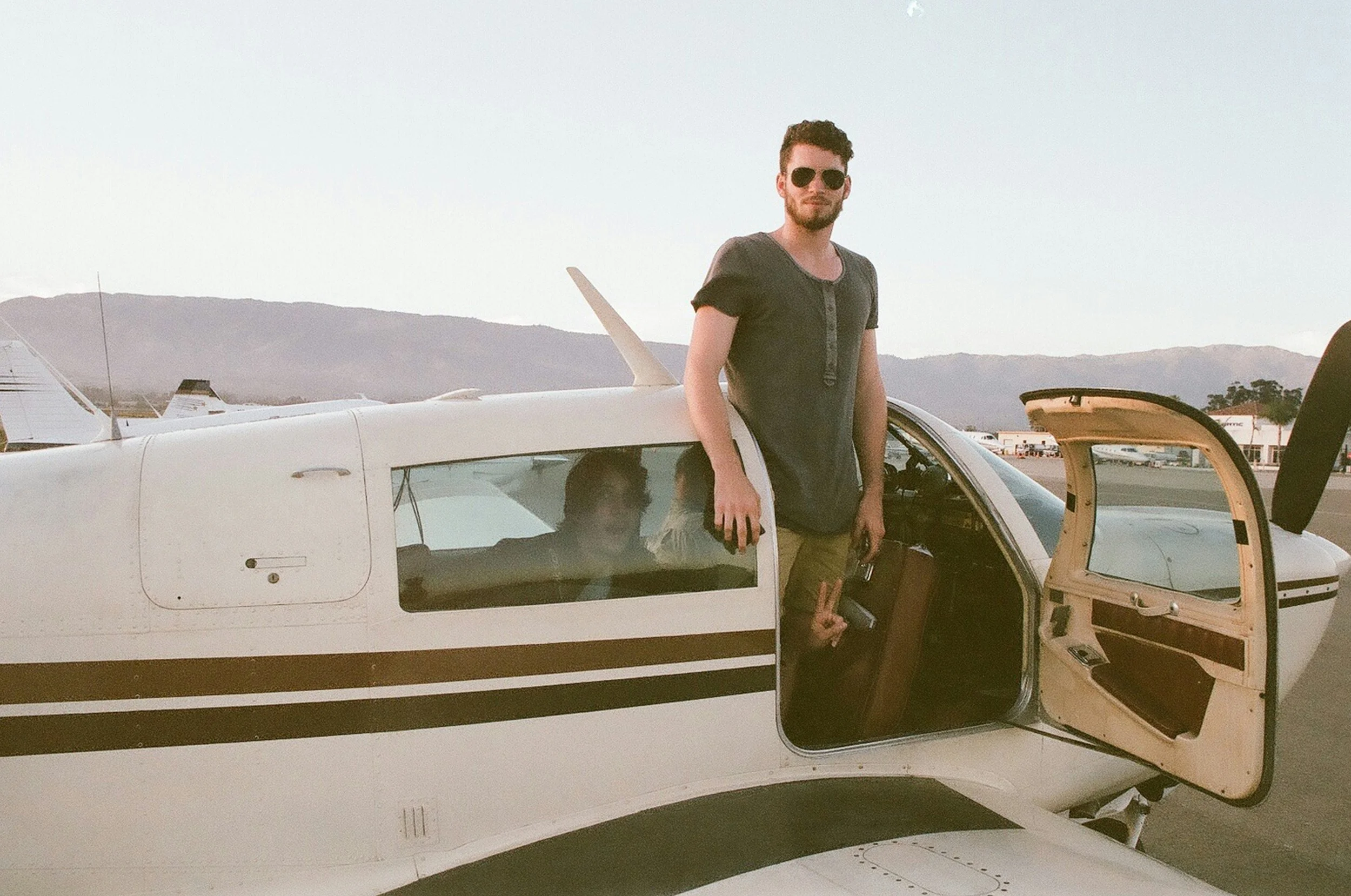 A man with sunglasses and a beard standing next to a small private airplane at an airfield, with mountains in the background.
