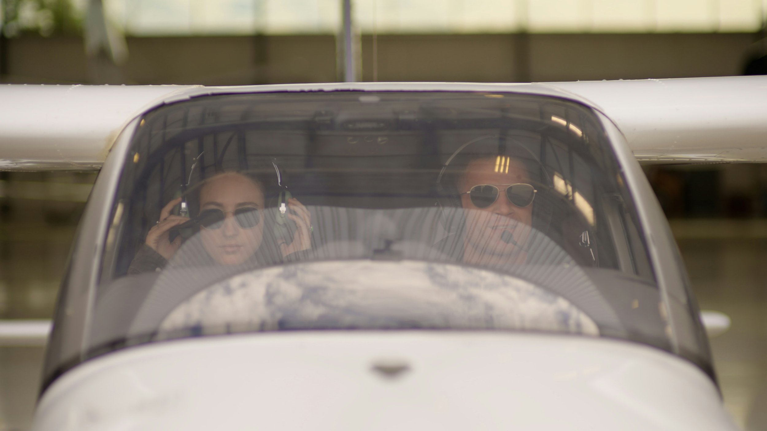 Two people in vintage airplane cockpit, one woman adjusting headphones and one man wearing sunglasses, both looking ahead.