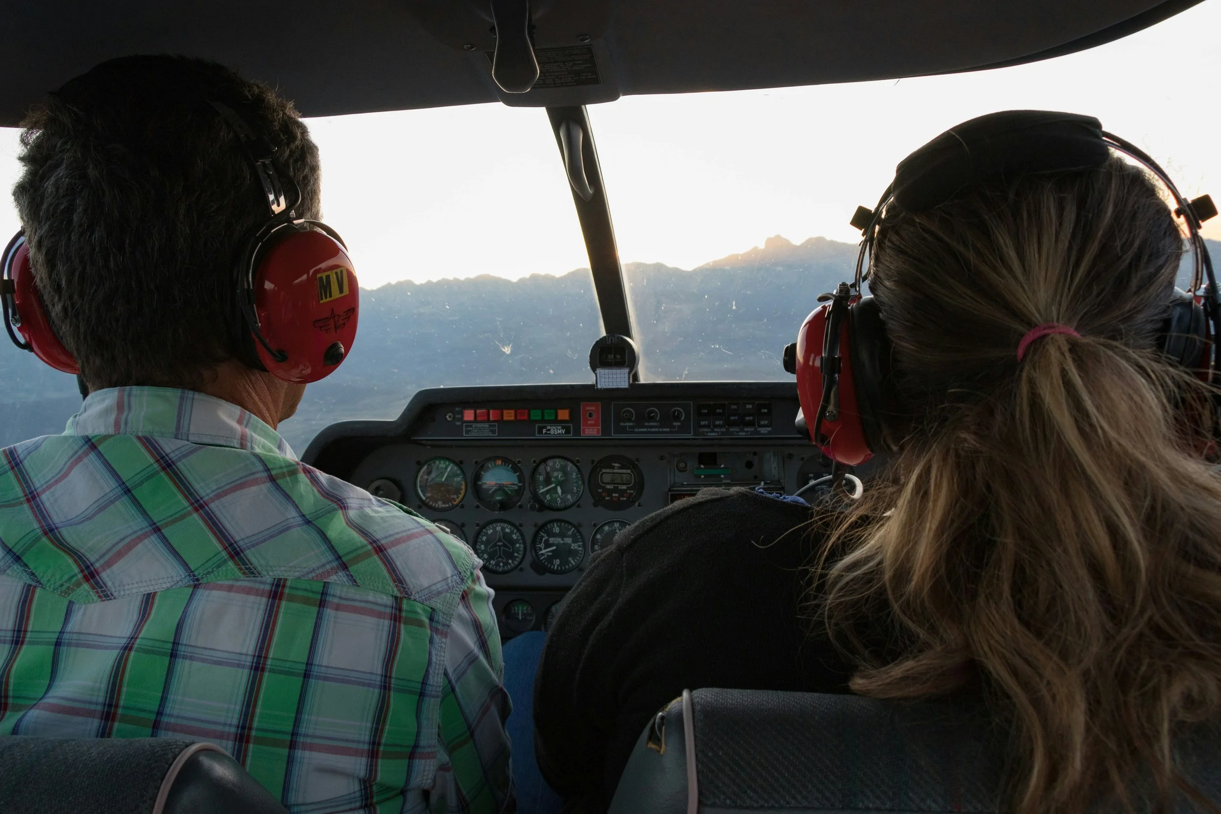 The interior of an airplane cockpit with two pilots, one man and one woman, flying over mountains at sunset. Both pilots are wearing headsets with microphones.