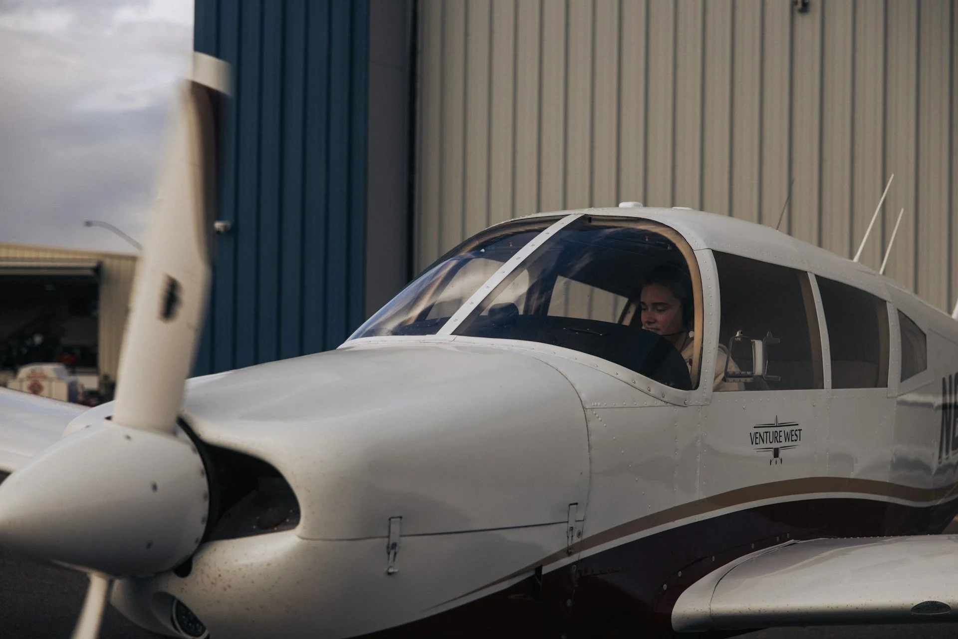 Close-up of a small white aircraft with a person inside the cockpit, parked near a building with vertical metal siding.