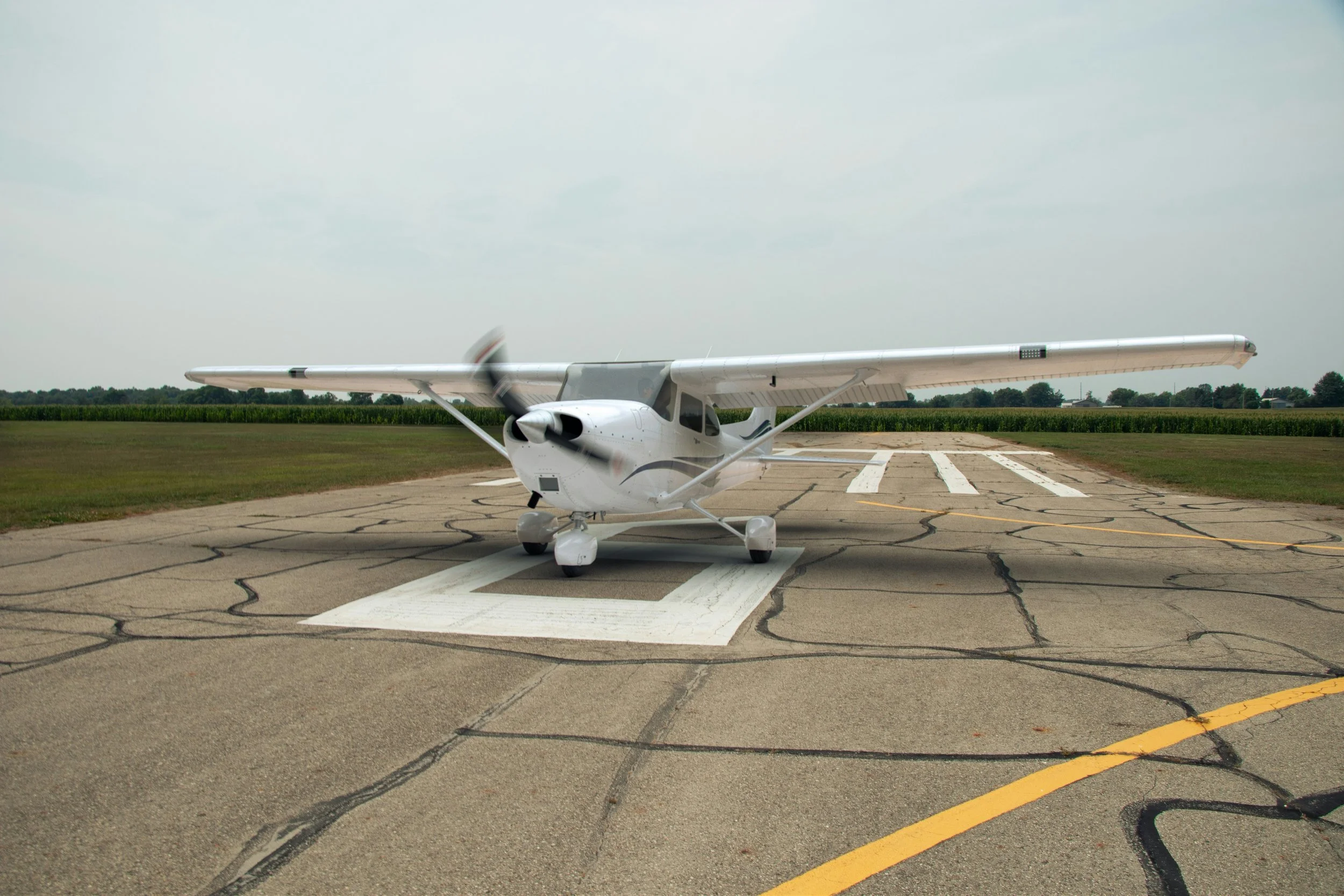 A small white airplane on a runway with green fields and trees in the background, under a cloudy sky.