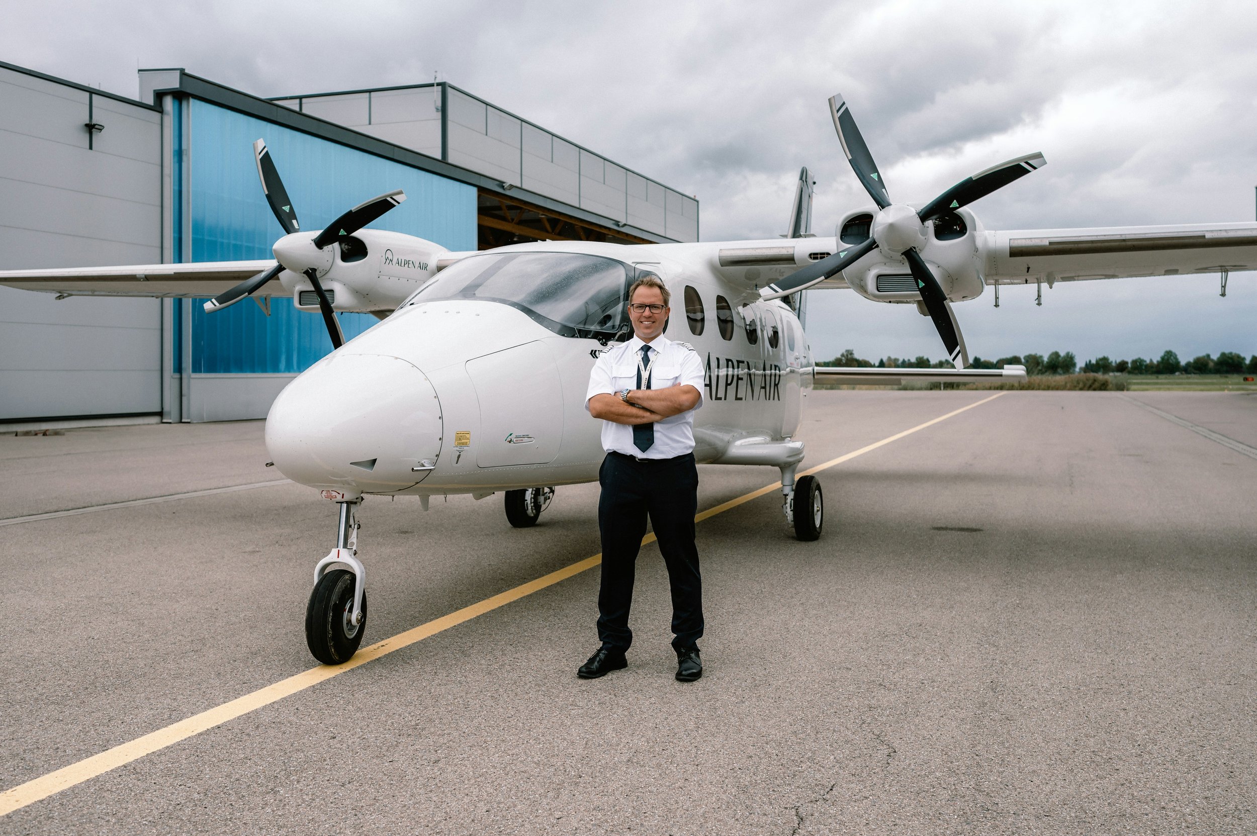 A man in a pilot uniform standing in front of a twin-engine airplane with propellers at an airport.
