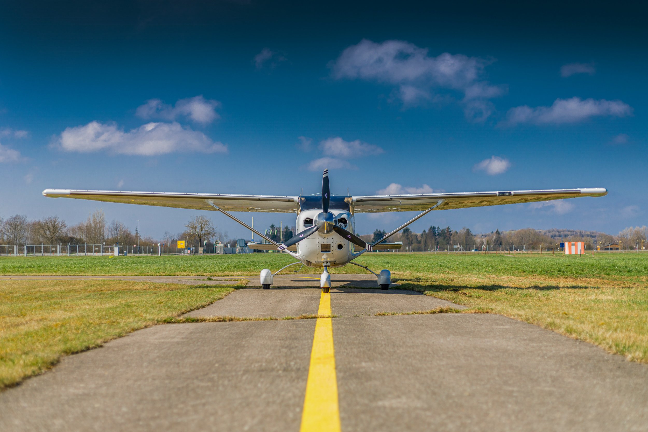 Small white airplane parked on the tarmac of an airport with a yellow line in front of it, surrounded by grass, with trees and a fence in the background under a partly cloudy sky.