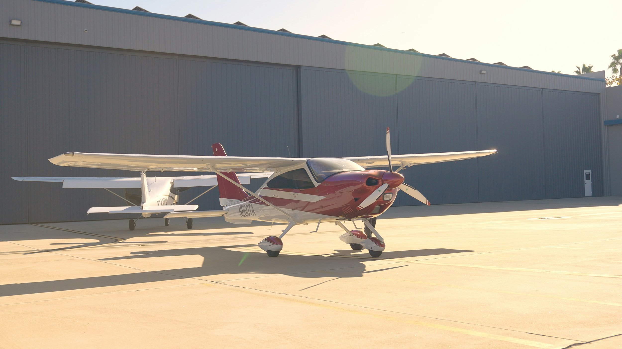 Small red and white airplane parked on a tarmac in front of a large grey hangar building with a garage door and a small access door.