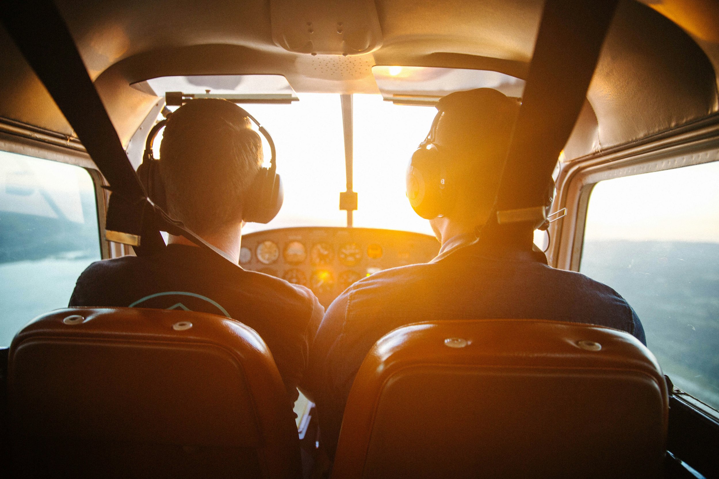 The interior of an airplane cockpit with two pilots wearing headsets, with sunlight streaming in through the windows.