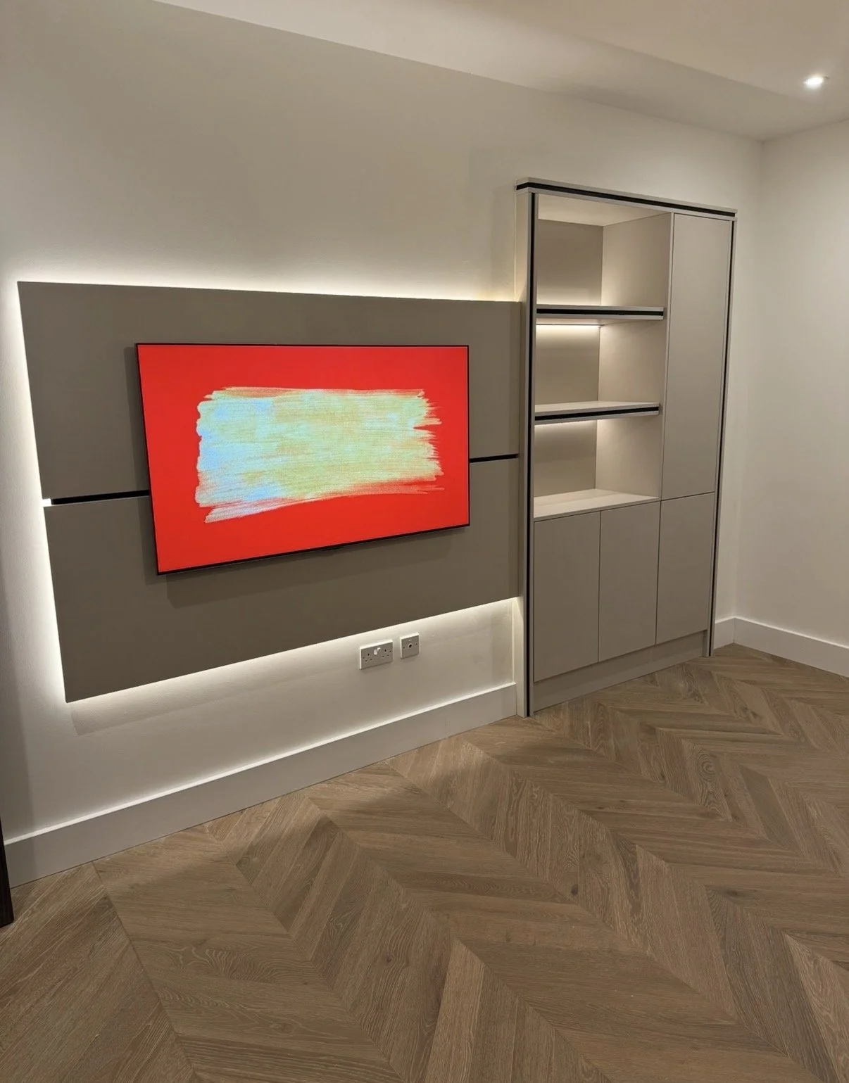 Modern interior with a flat-screen TV mounted on a beige wall, displaying abstract art with red, yellow, light blue, and white colors. Next to the TV is a built-in cabinet with open shelves, closed compartments, and LED lighting. The floor is made of