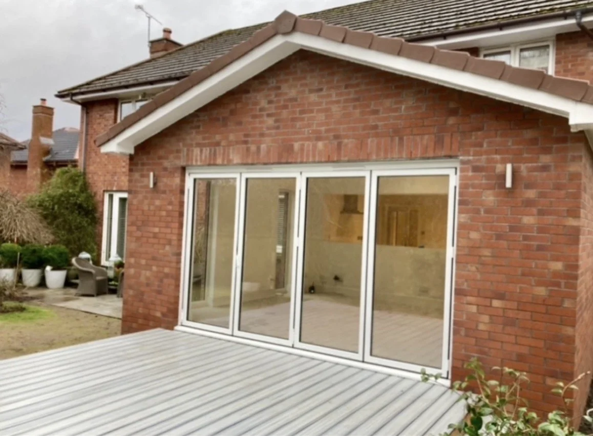 Brick house with large glass sliding doors leading to a wooden deck, in a suburban backyard with potted plants and garden furniture.