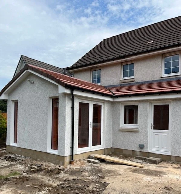 The back of a newly built two-story house with a small attached one-story extension. The extension has a pitched roof with red tiles, white stucco walls, and several windows and a door. The ground around the house is unfinished, with dirt and constru