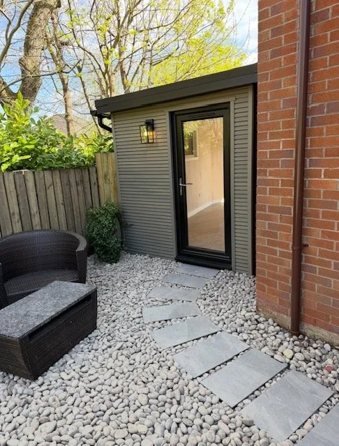 Small backyard patio with stepping stones, white gravel, a black outdoor chair, a small bush, a gray shed with a glass door, a brick wall, a wood fence, and trees in the background.