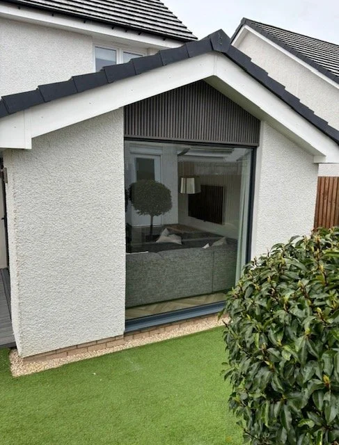 View of a house's exterior with a large window, seen through which a living room with a TV and a decorative tree inside. The house has white textured walls, a dark roof, and a small lawn area with green grass outside.
