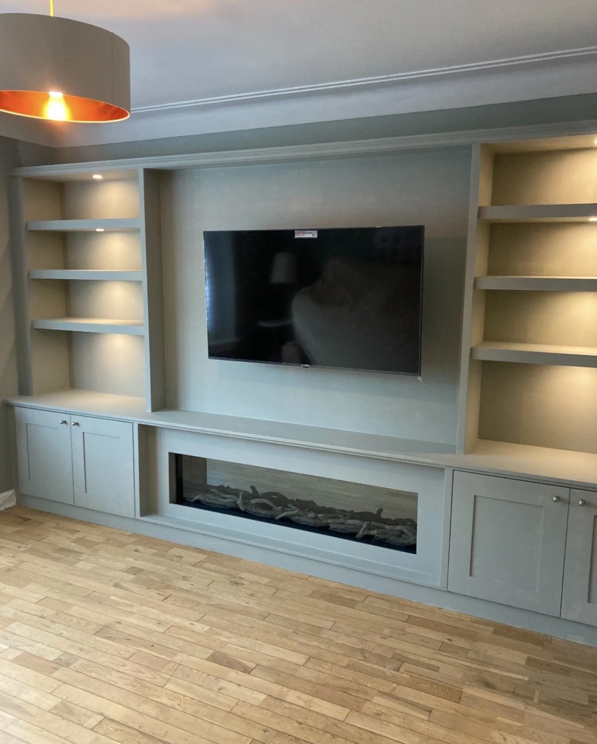 Living room entertainment center with built-in shelves, a mounted flat-screen TV, and a fireplace below, in a room with wooden flooring and a ceiling light.
