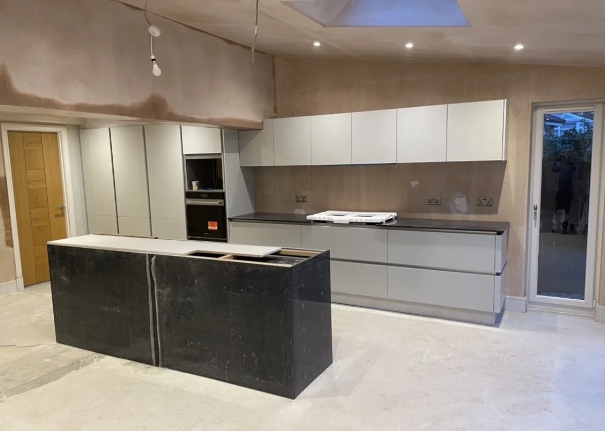 Unfinished modern kitchen with gray cabinets, black countertop, and an island with black marble surface, with a door and sliding glass door, and exposed ceiling wiring.