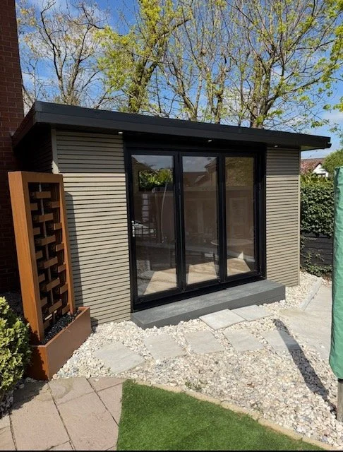 Small modern backyard building with sliding glass doors, surrounded by gravel and decorative paving stones, with trees and shrubs in the background.