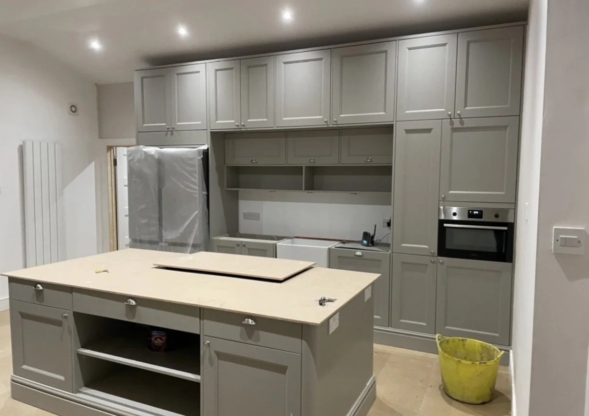 Kitchen under renovation with grey cabinetry, an island, and appliances covered in plastic, with a yellow bucket on the floor.