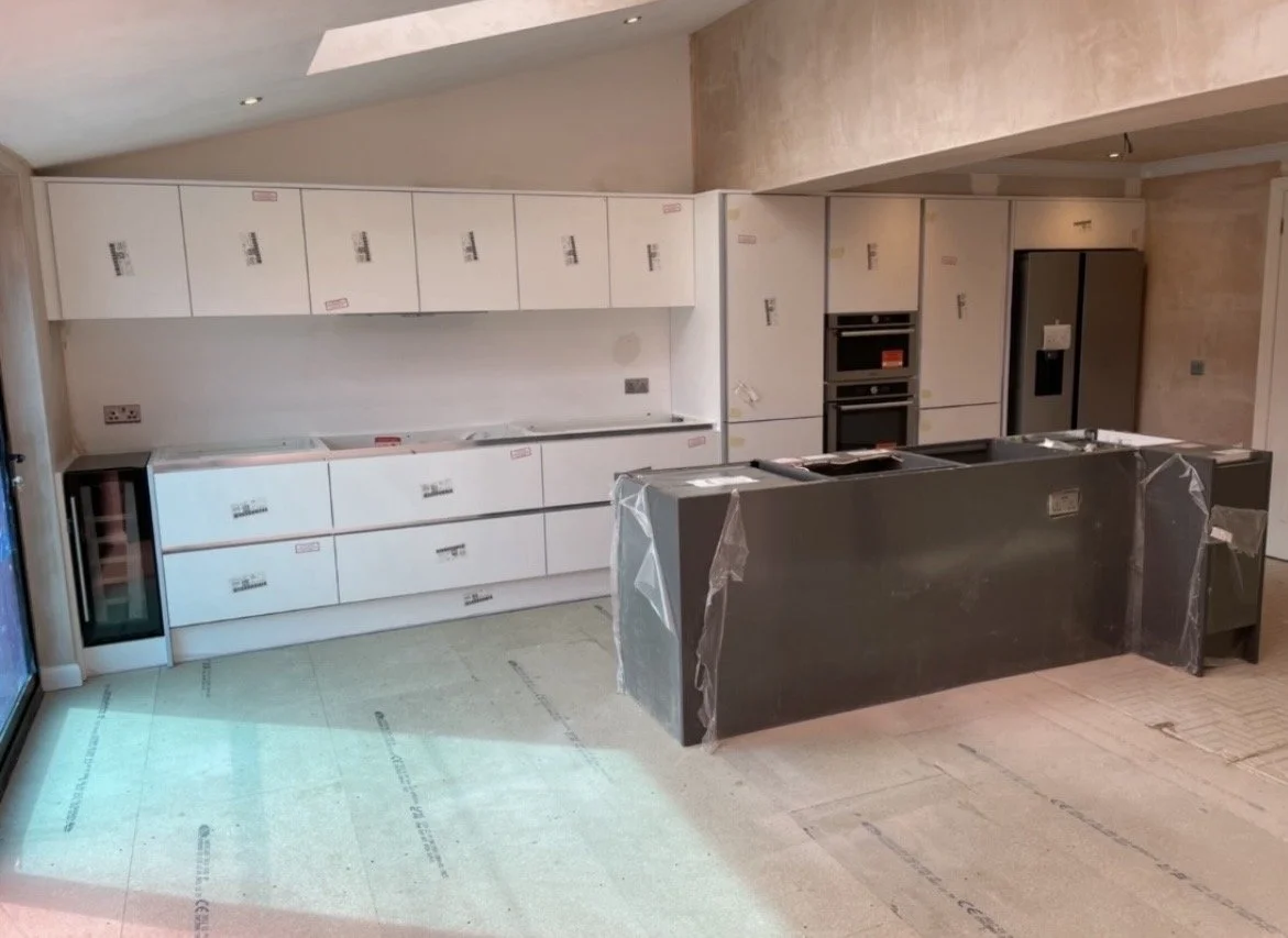 Kitchen under construction with white cabinets, a black kitchen island wrapped in protective plastic, and built-in appliances including double ovens and a large refrigerator.